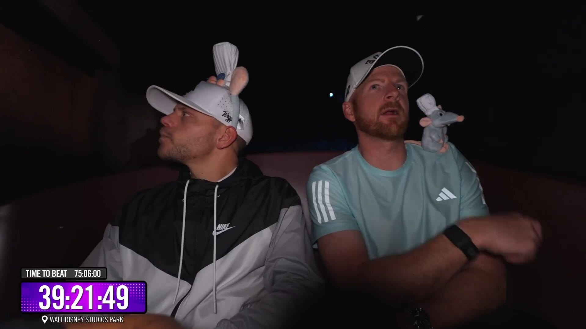 Two men in baseball caps with Remy plush toys ride a dark Disney Park attraction; VIDEO timer graphic in corner.