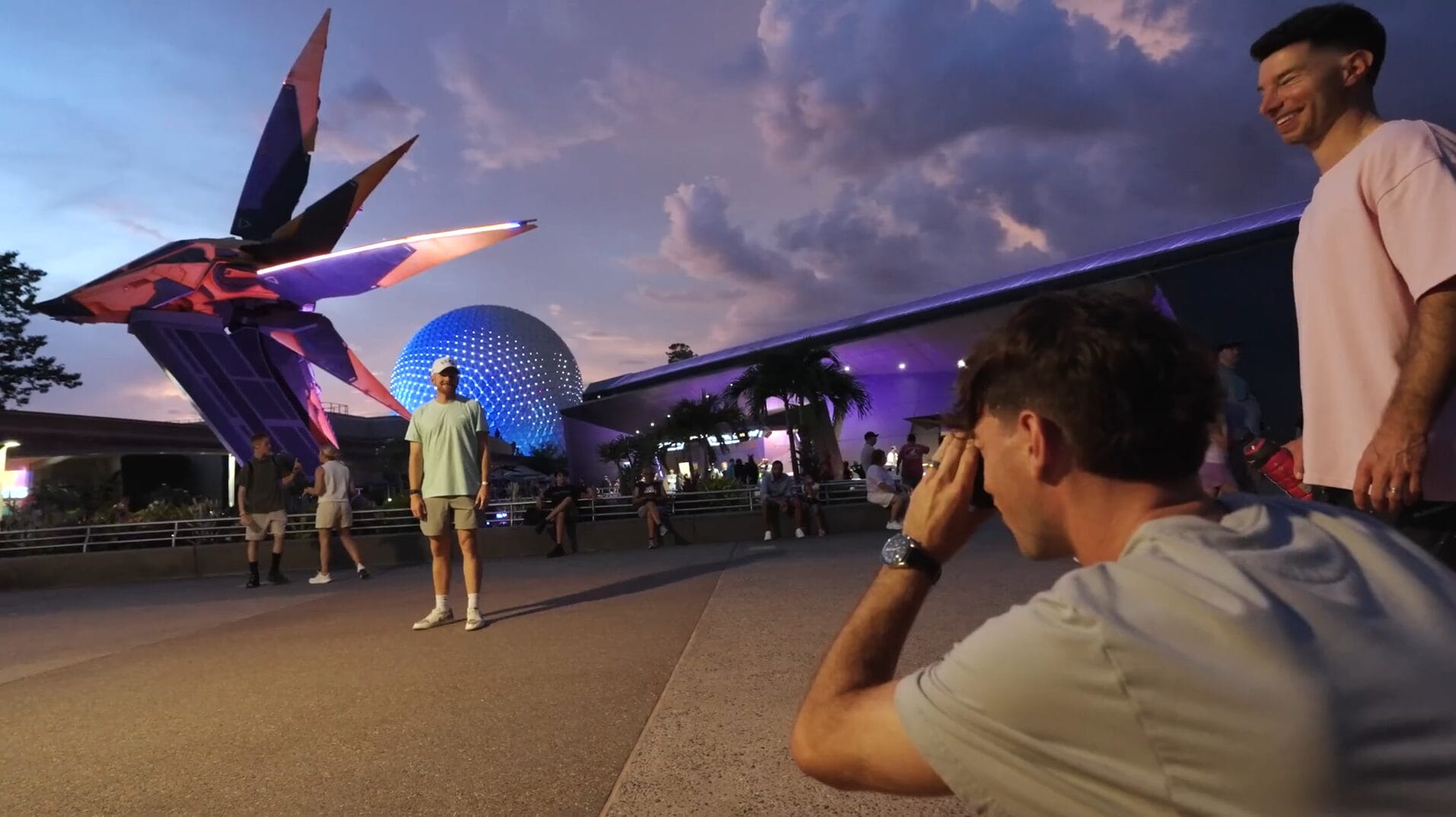 A man kneels to take a photo of another man posing near a large spaceship statue at sunset in a Disney Park.
