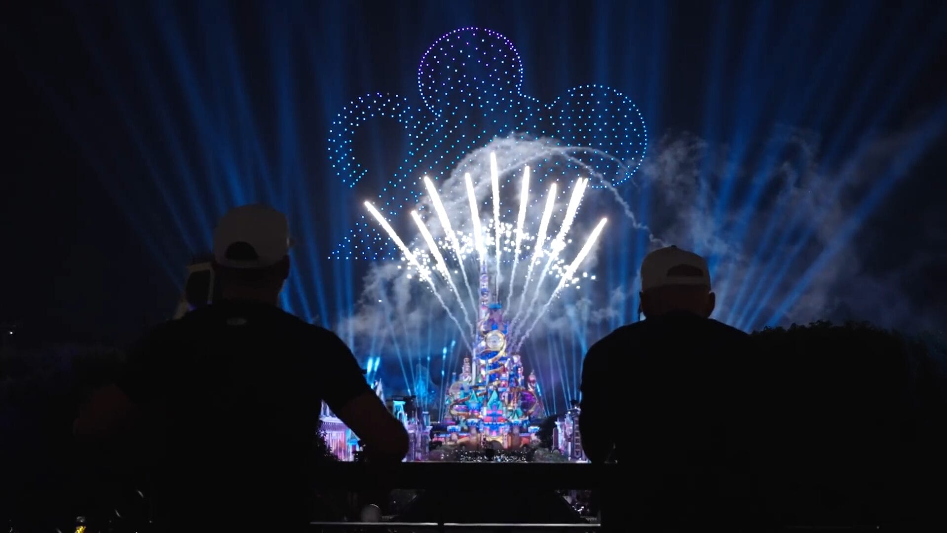 Two people watch fireworks and a Dude Perfect drone show above a brightly lit castle at Disney Park.