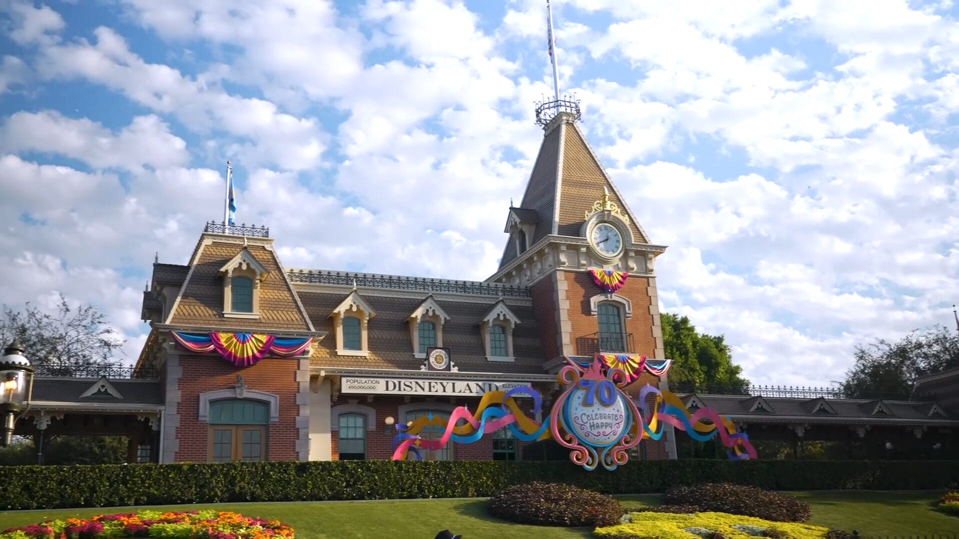 Disney Park entrance with Dude Perfect 70th anniversary decorations and vibrant flowers under a partly cloudy sky.