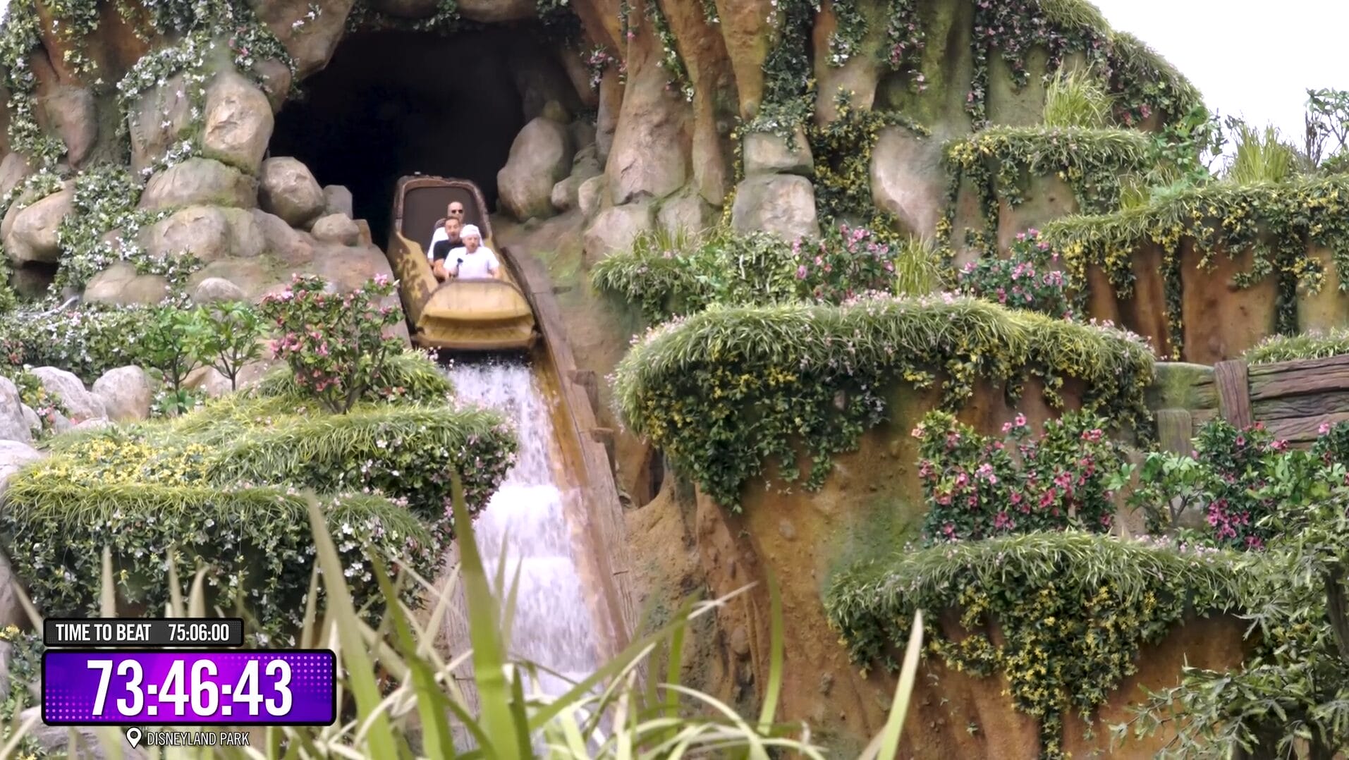 A log flume ride emerges from a cave, like a Disney Park adventure, descending a splashy drop amid rocks and greenery.