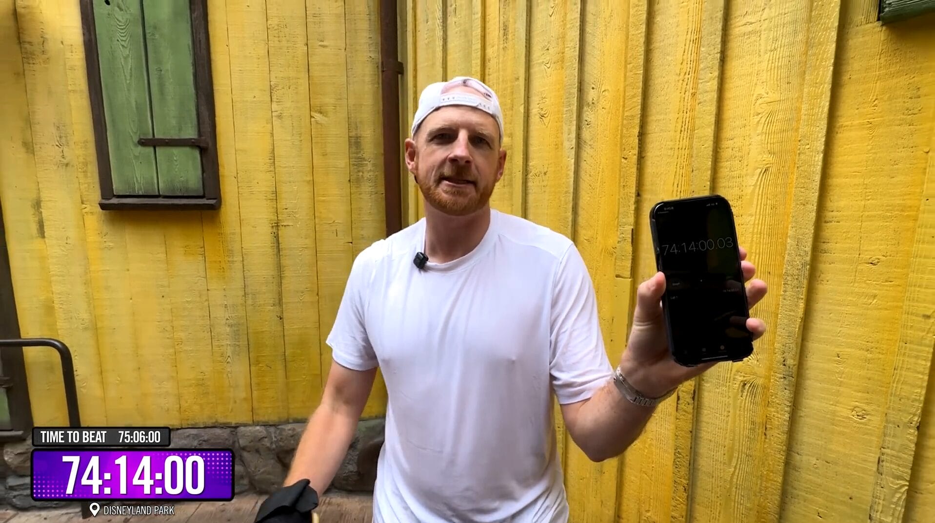 A man in a white shirt holds up a phone with a timer, recording a video in front of a yellow wooden wall.
