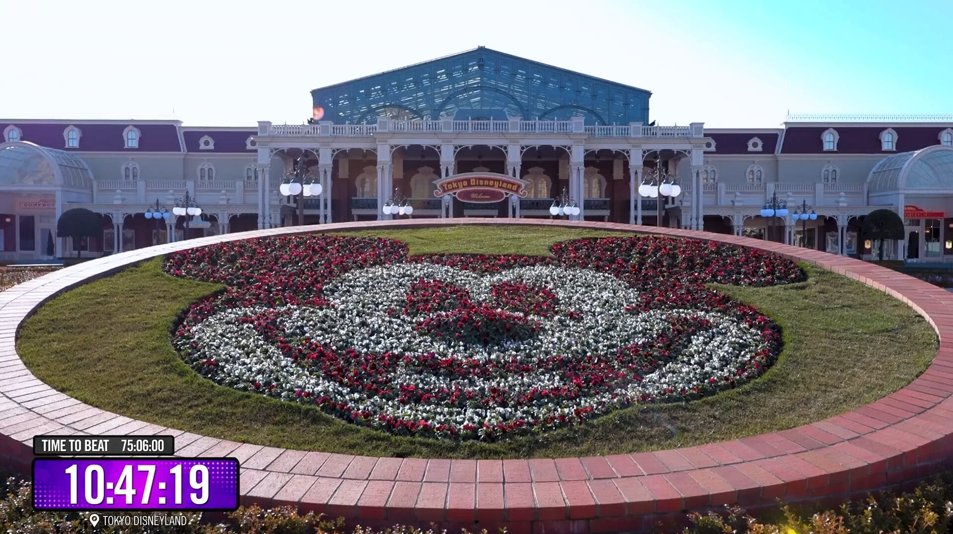 A flower bed shaped like a cartoon mouse face in front of a grand Disney Park building, timer at bottom left in video.