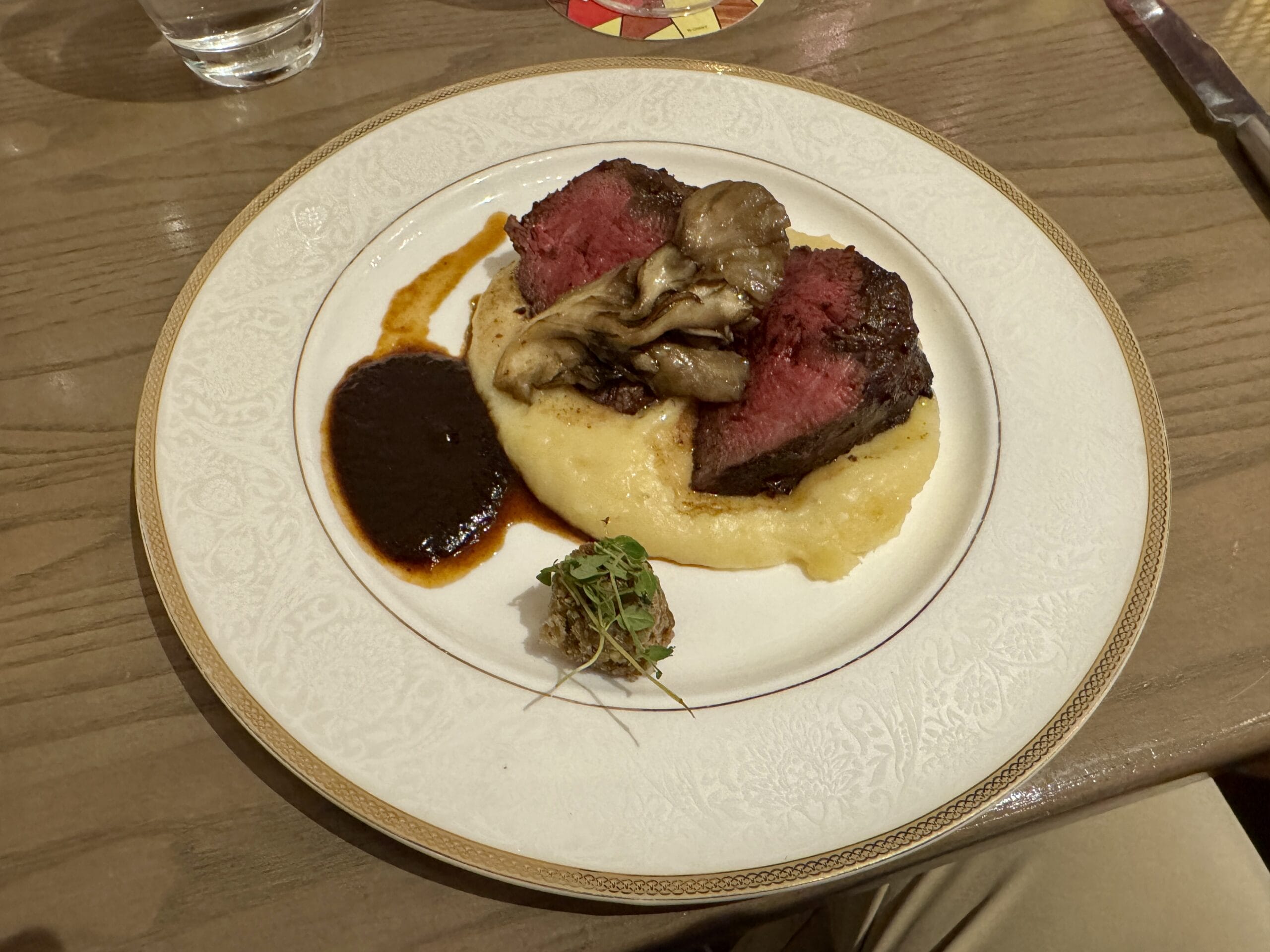 A plate of sliced steak tapas on mashed potatoes, with mushrooms, sauce, and a garnish sits on a Toledo-style wooden table.