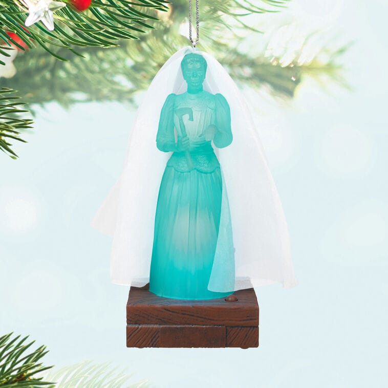 A blue translucent bride Hallmark ornament with a white veil, reminiscent of the Haunted Mansion at Disney parks, hangs on a tree.