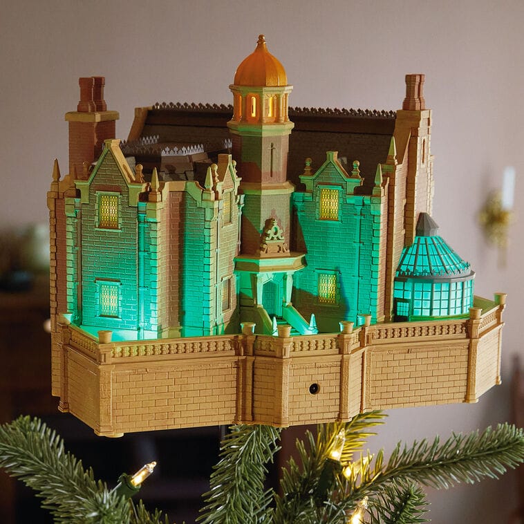 A Hallmark model of a mansion styled like a Disney castle, glowing green and yellow, crowns a Christmas tree, evoking Cinderella Castle.