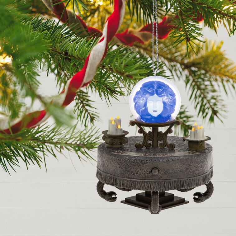 A Haunted Mansion Disney Keepsake Ornament of a seance table with glowing blue face, hanging on a Christmas tree branch.