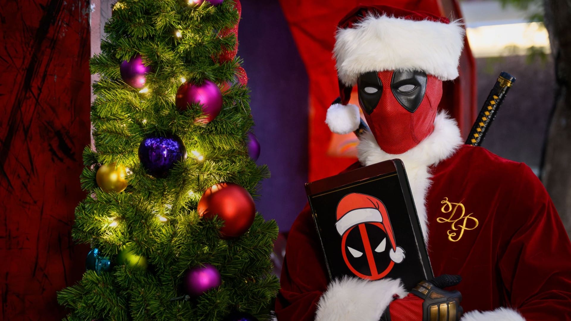 Deadpool in a Santa suit poses by a decorated tree, holding a festive Daily Recap book—Disney park magic meets holiday cheer.