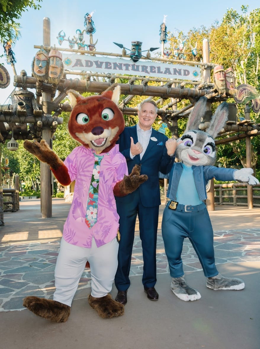 Two in animal costumes and a suited man pose at a Disney Adventureland Meet & Greet—no castle like WDW or other Disneyland parks.