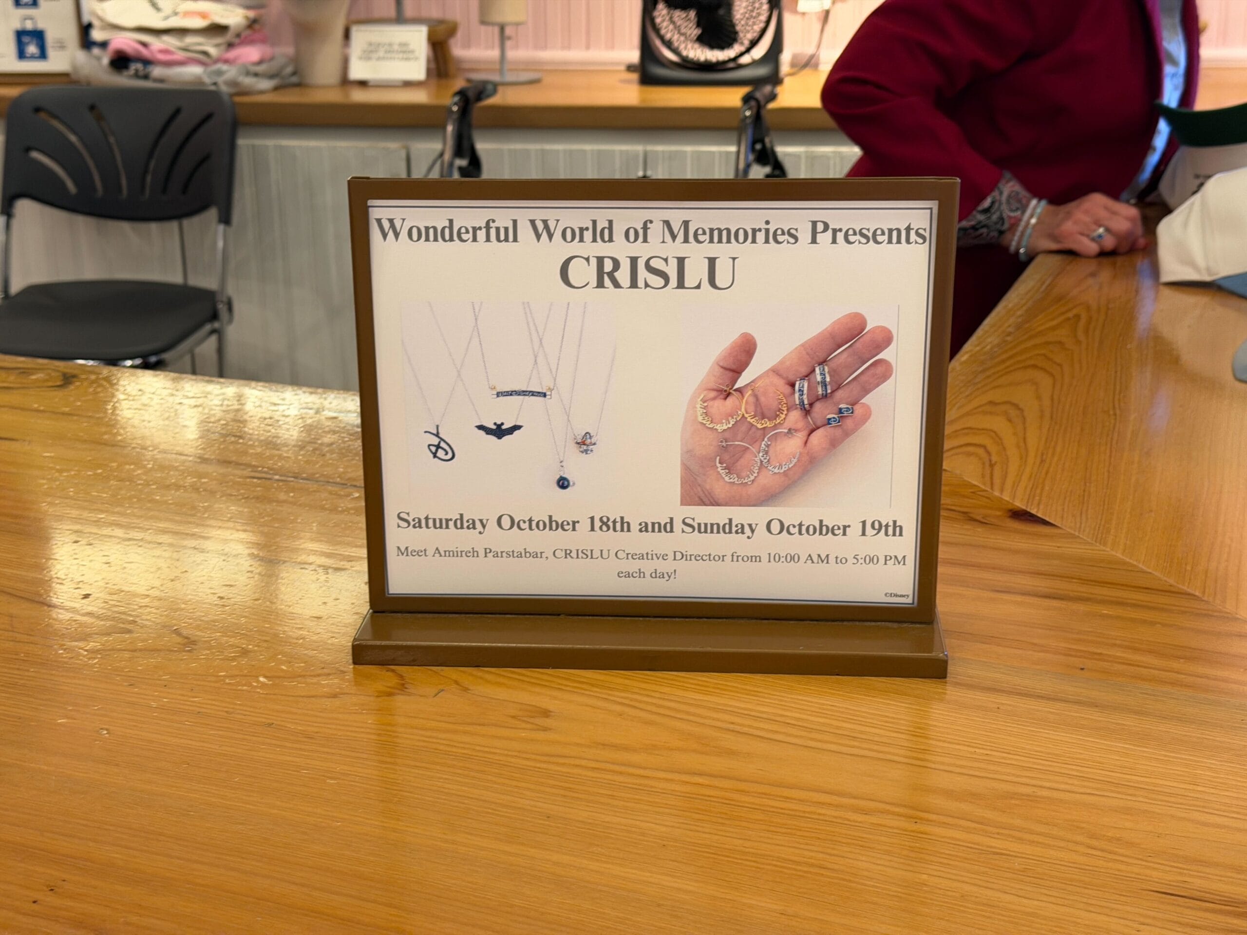Sign on a counter in a Disney park promotes CRISLU jewelry Halloween Trunk Show, Oct 18–19, with castle-themed necklaces and bracelets.