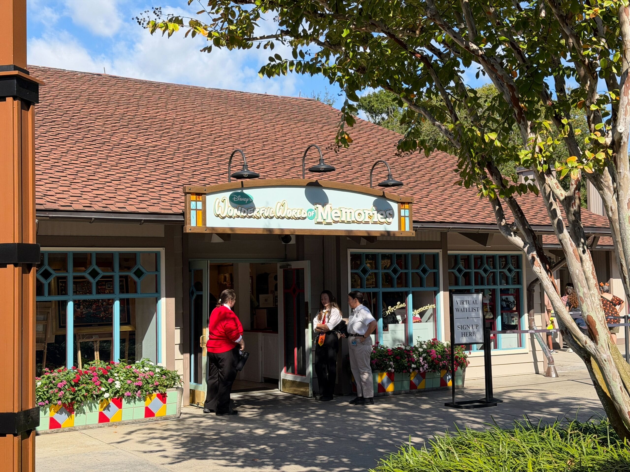 Three guests chat outside "Wonderful World of Memories" at Walt Disney World, flowers and trees nearby on a bright sunny day.