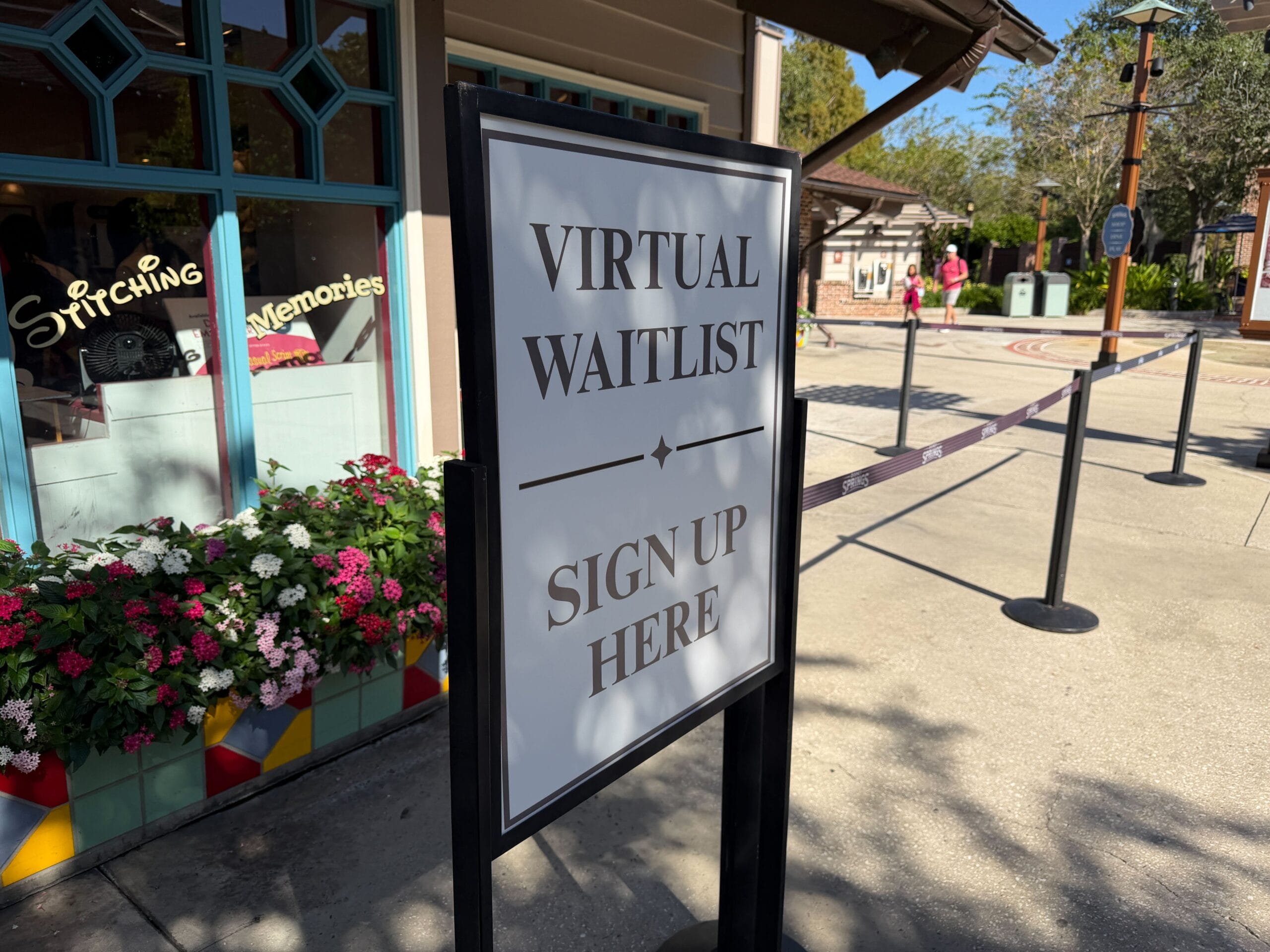 A "Virtual Waitlist Sign Up Here" sign stands by flowers and an empty walkway at Walt Disney World, no castle in view.