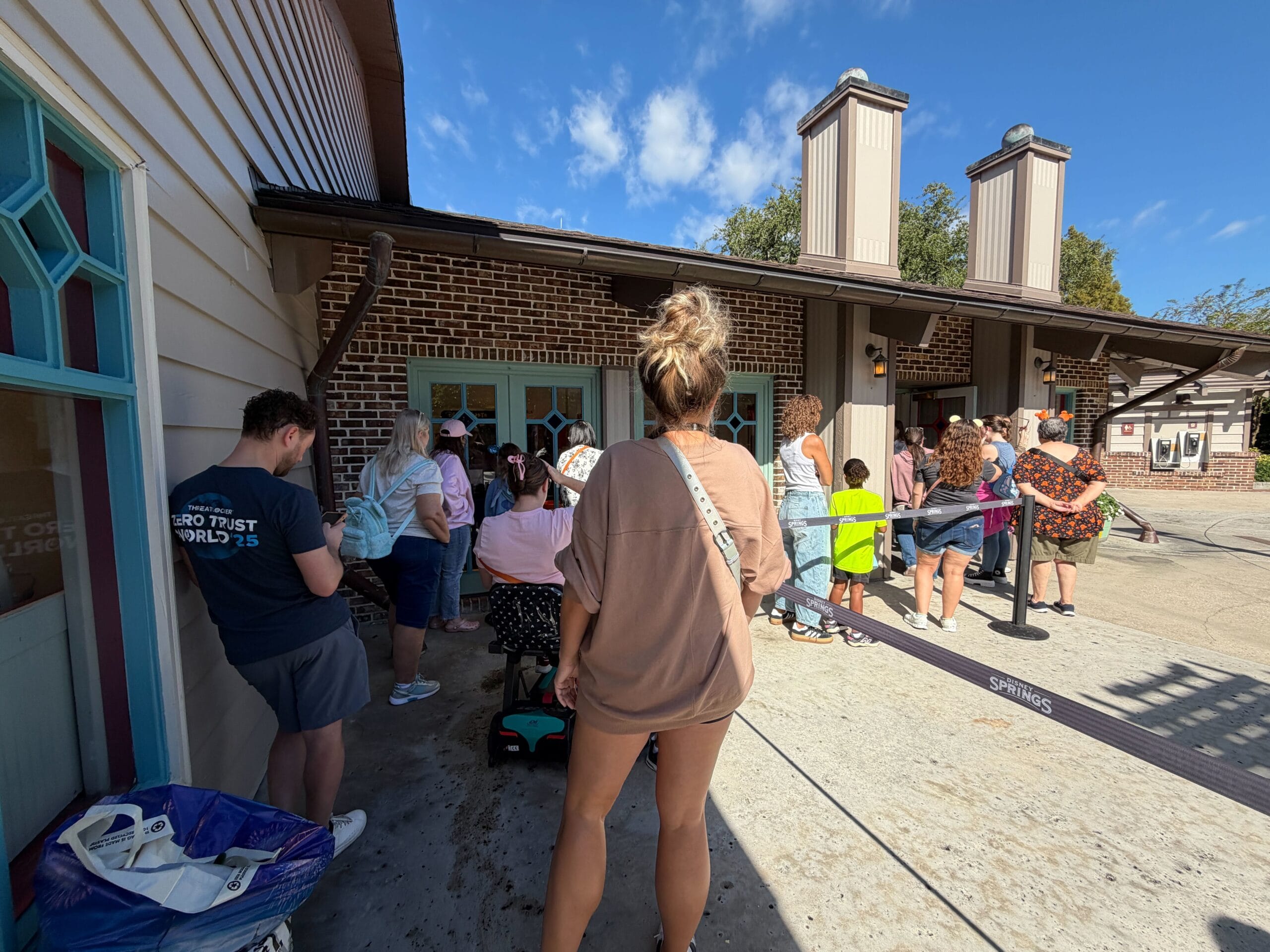 Guests wait in line outside a building at a Disney park on a sunny day, anticipating the Halloween Trunk Show by the entrance.