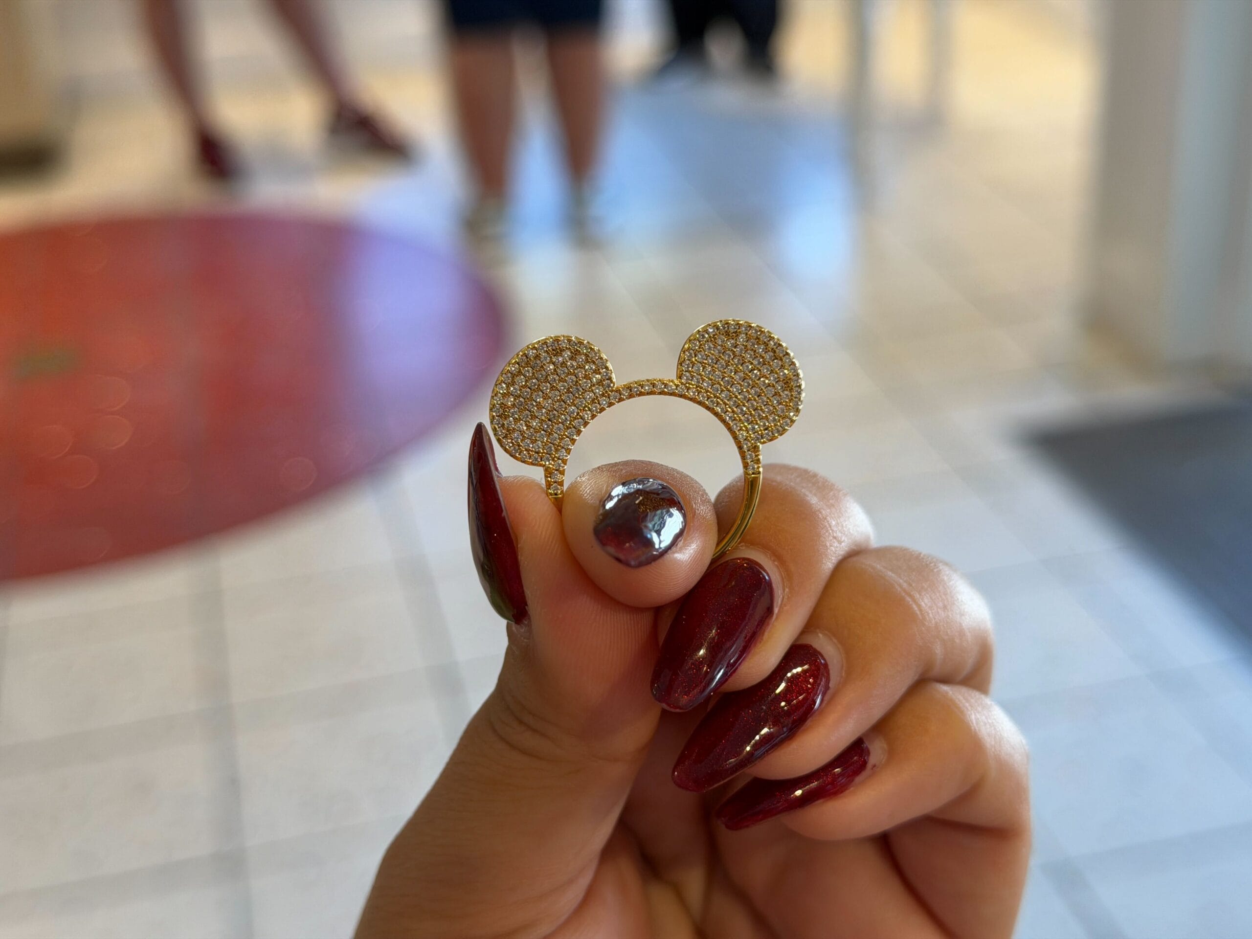 A hand with red nails holds a gold CRISLU Mickey Mouse ring with rhinestones, reminiscent of Disney Parks merchandise, indoors on tile.