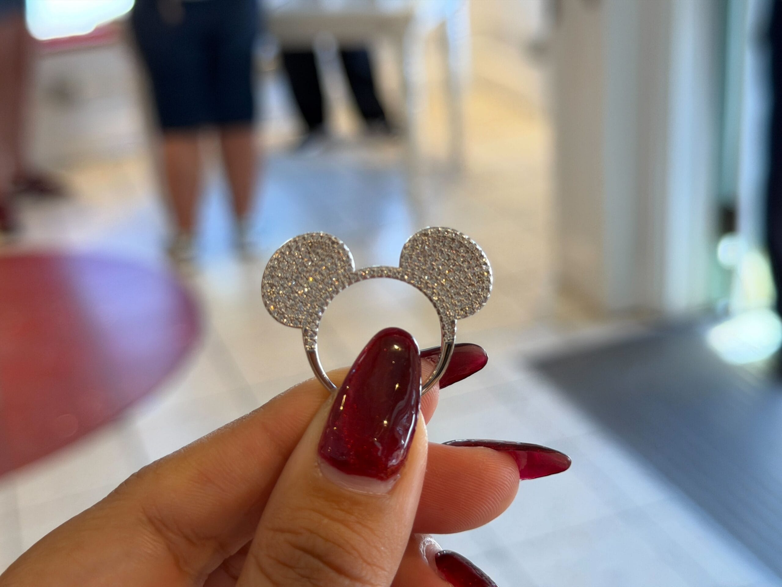 A hand with red nails displays a glittering CRISLU Mickey Mouse ring at a Walt Disney World Halloween Trunk Show, indoors.