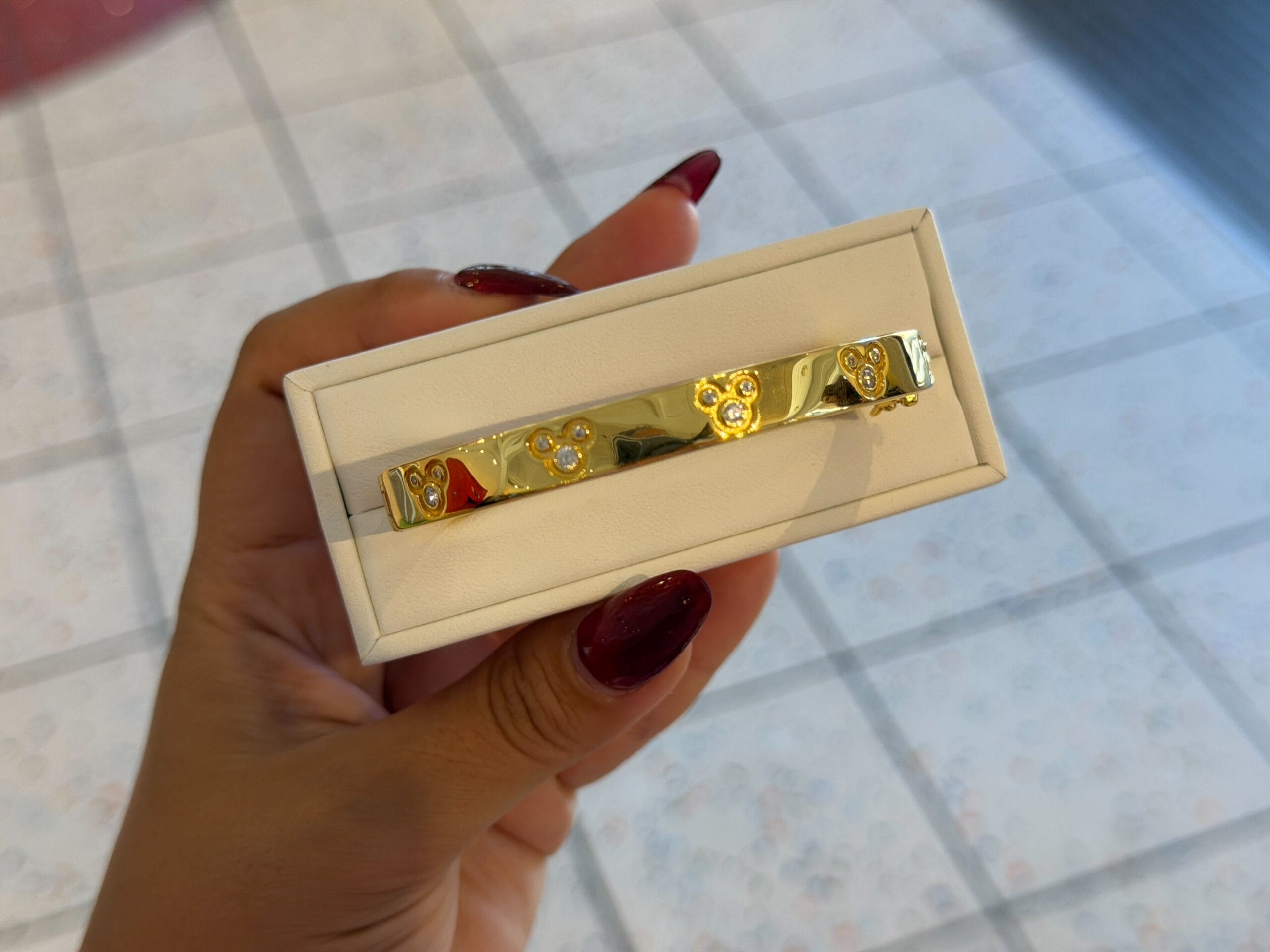 A hand with red nails holds a gold CRISLU bracelet with floral patterns in a white jewelry box, like those in Disney gift shops.