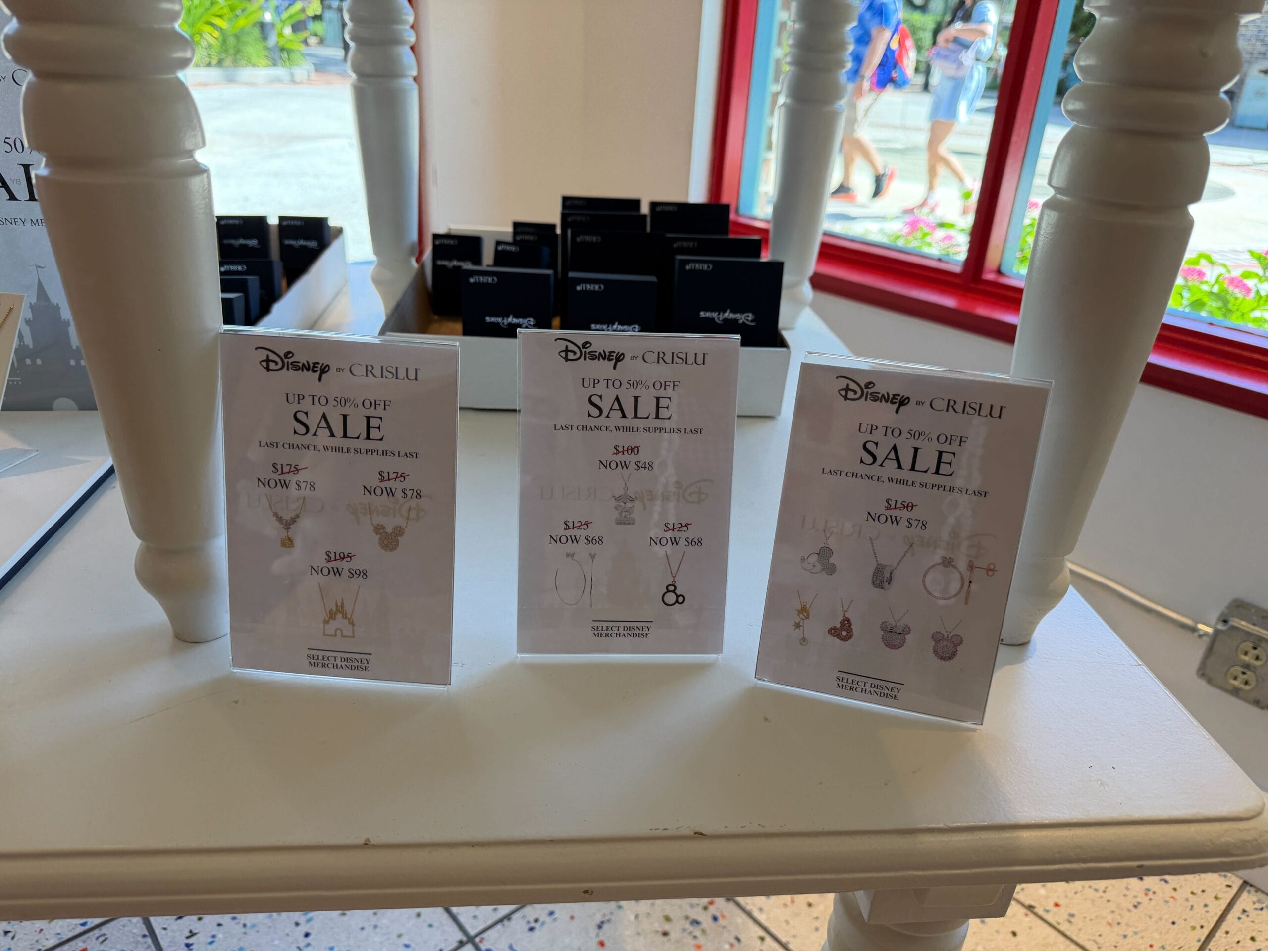 Three sale signs for Disney x CRISLU jewelry promote Halloween Trunk Show discounts on a white table at a Disney park store.