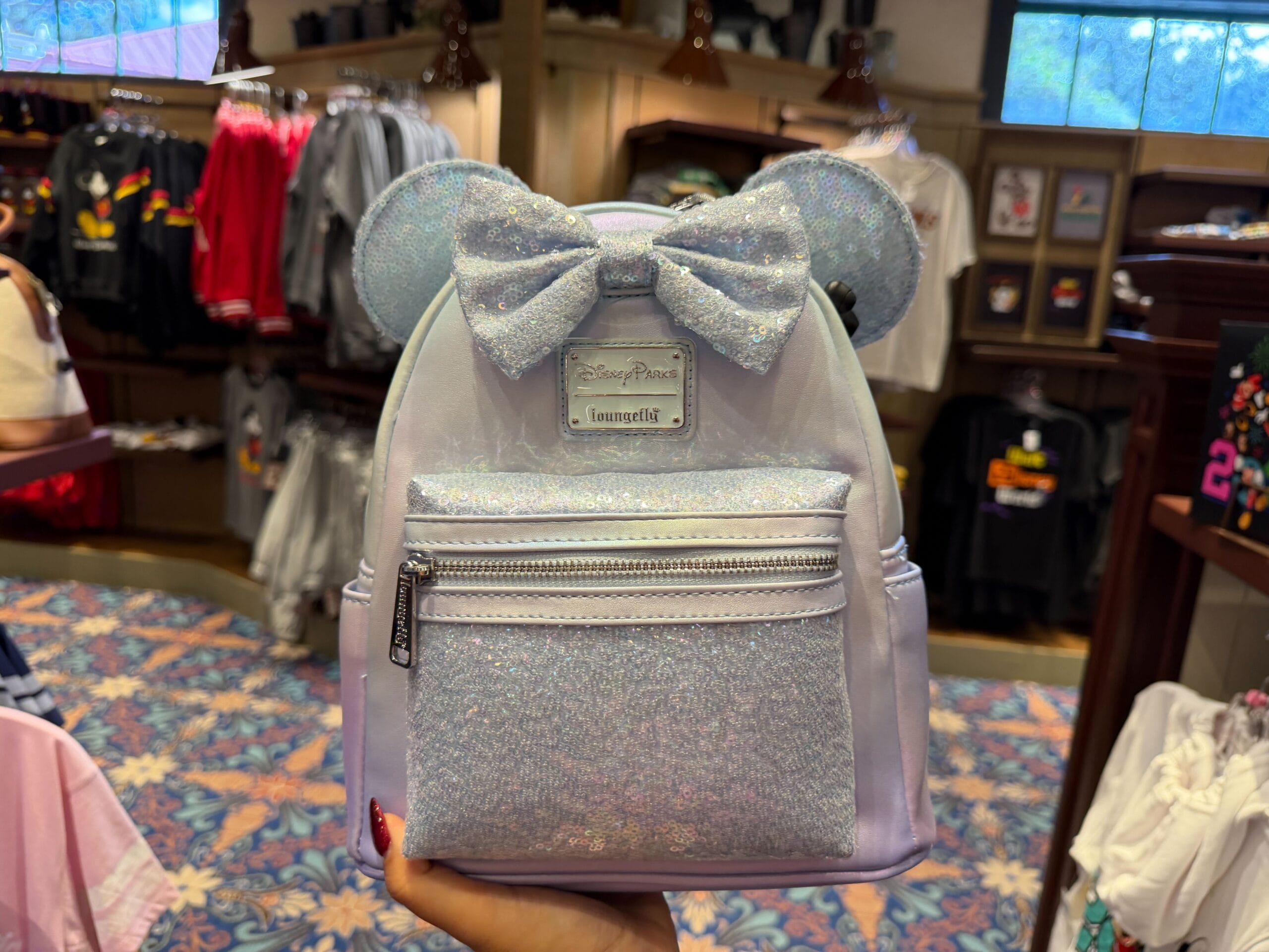 A sparkly silver Disney backpack with mouse ears and a bow is showcased in a park store, colorful Disney apparel behind it.