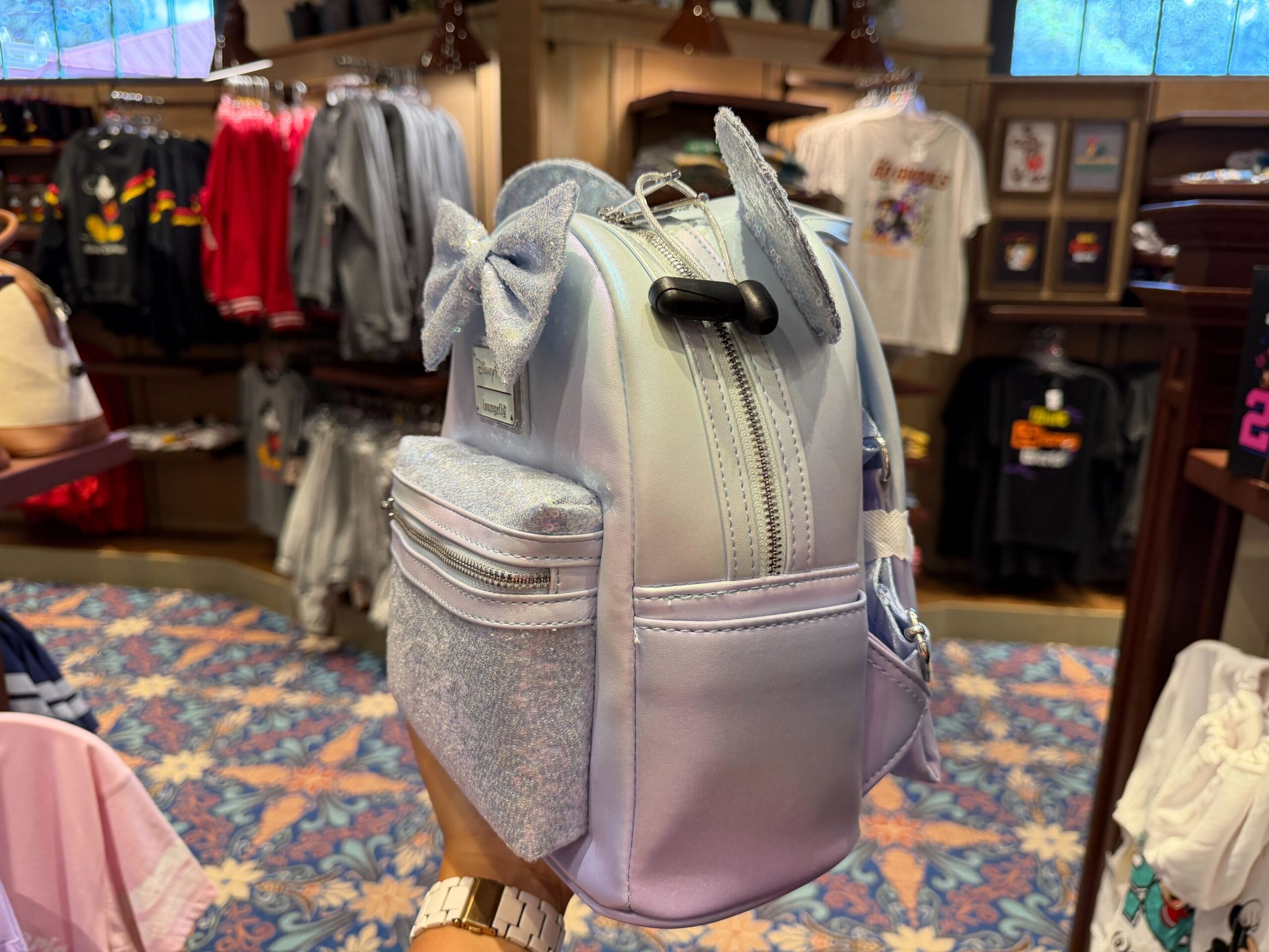 A guest holds a pastel purple mini backpack with bow and Mickey ears in a Disney Park shop with patterned carpet, no castle in sight.