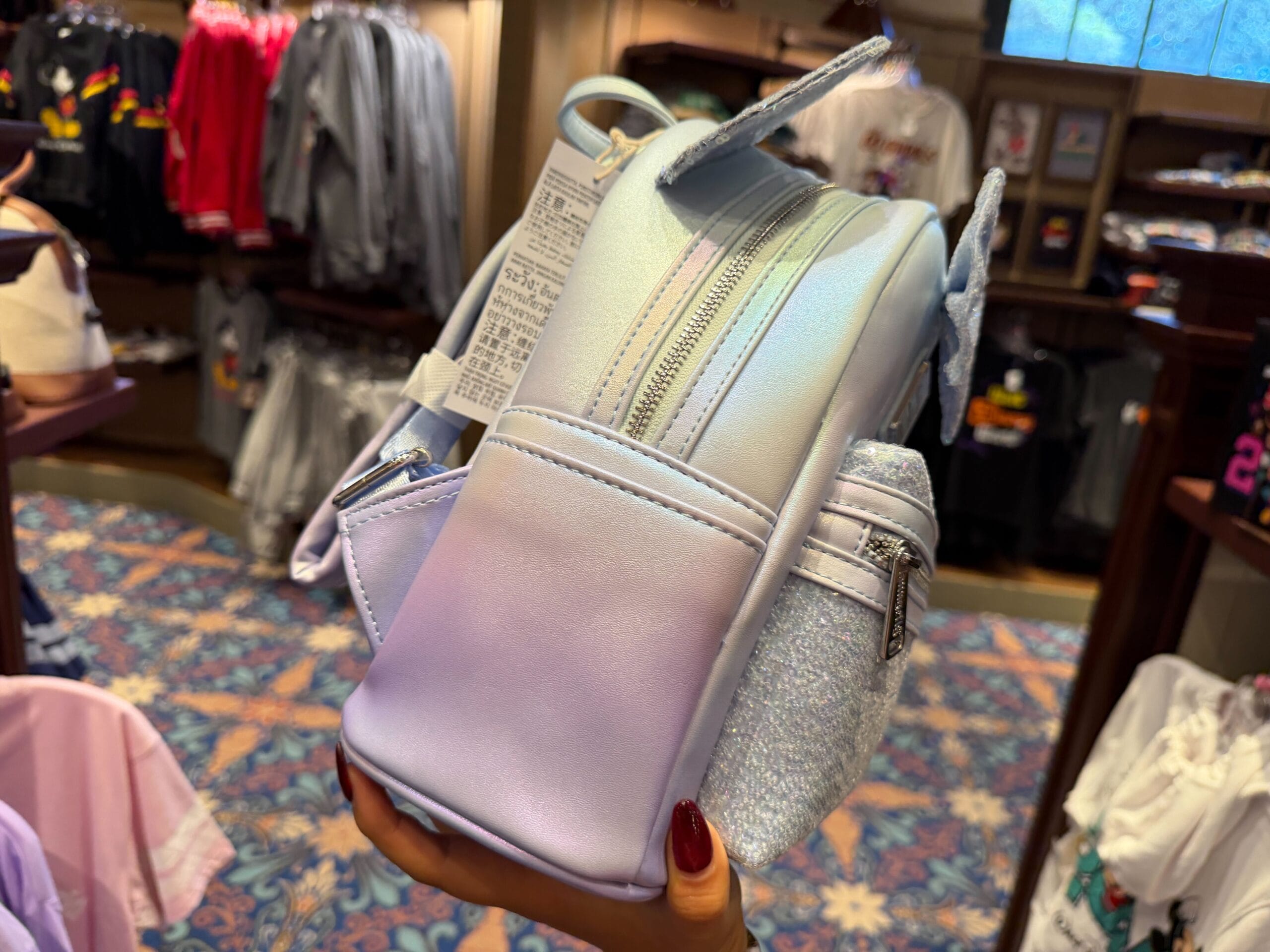 A hand holds a small iridescent pastel backpack with silver zippers, displayed inside a Disney park shop, reminiscent of park merch.