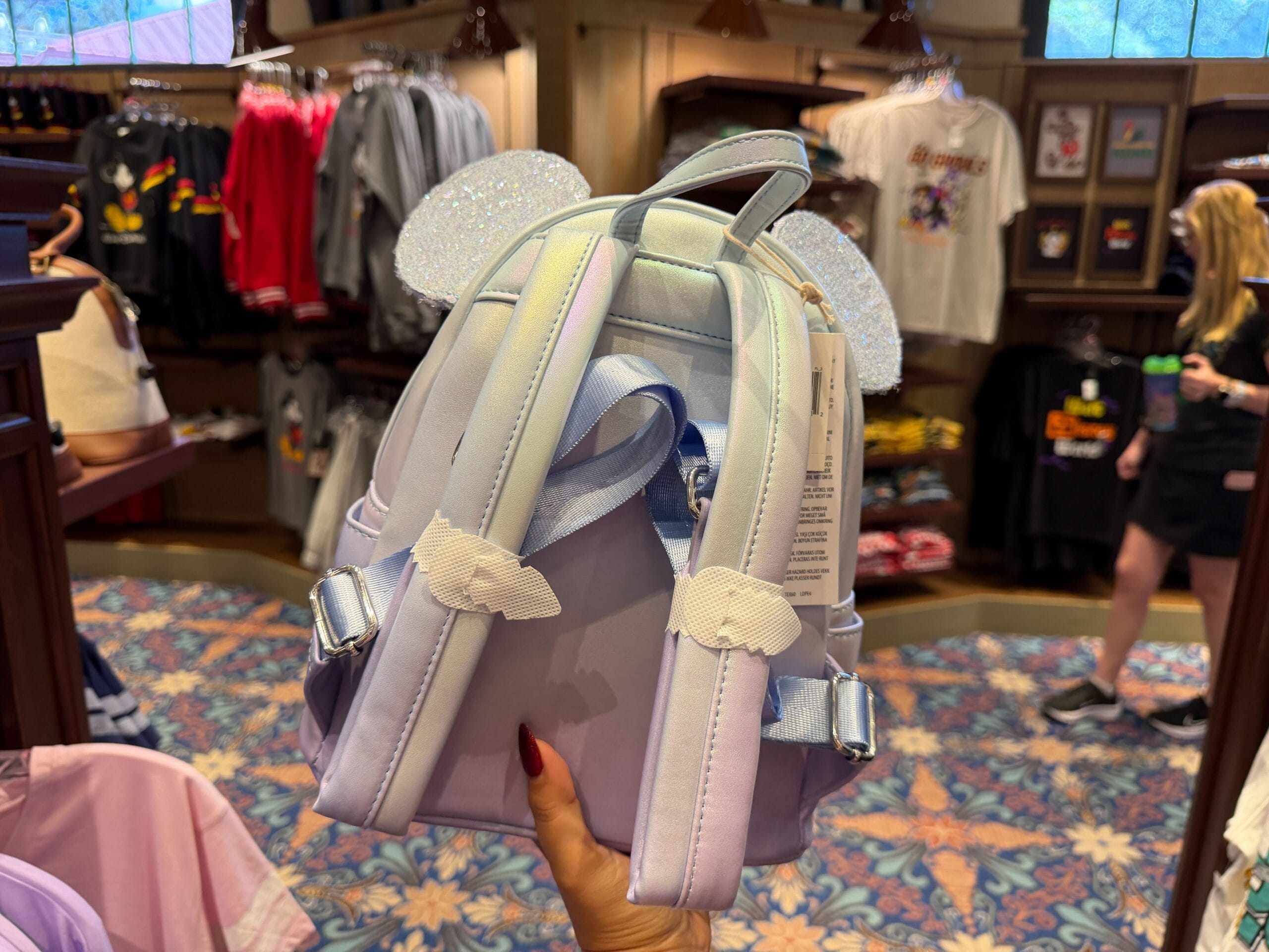 Guest holds a pastel backpack with sparkly Mickey ears and wings in a Disney park shop, surrounded by colorful shirts on display.
