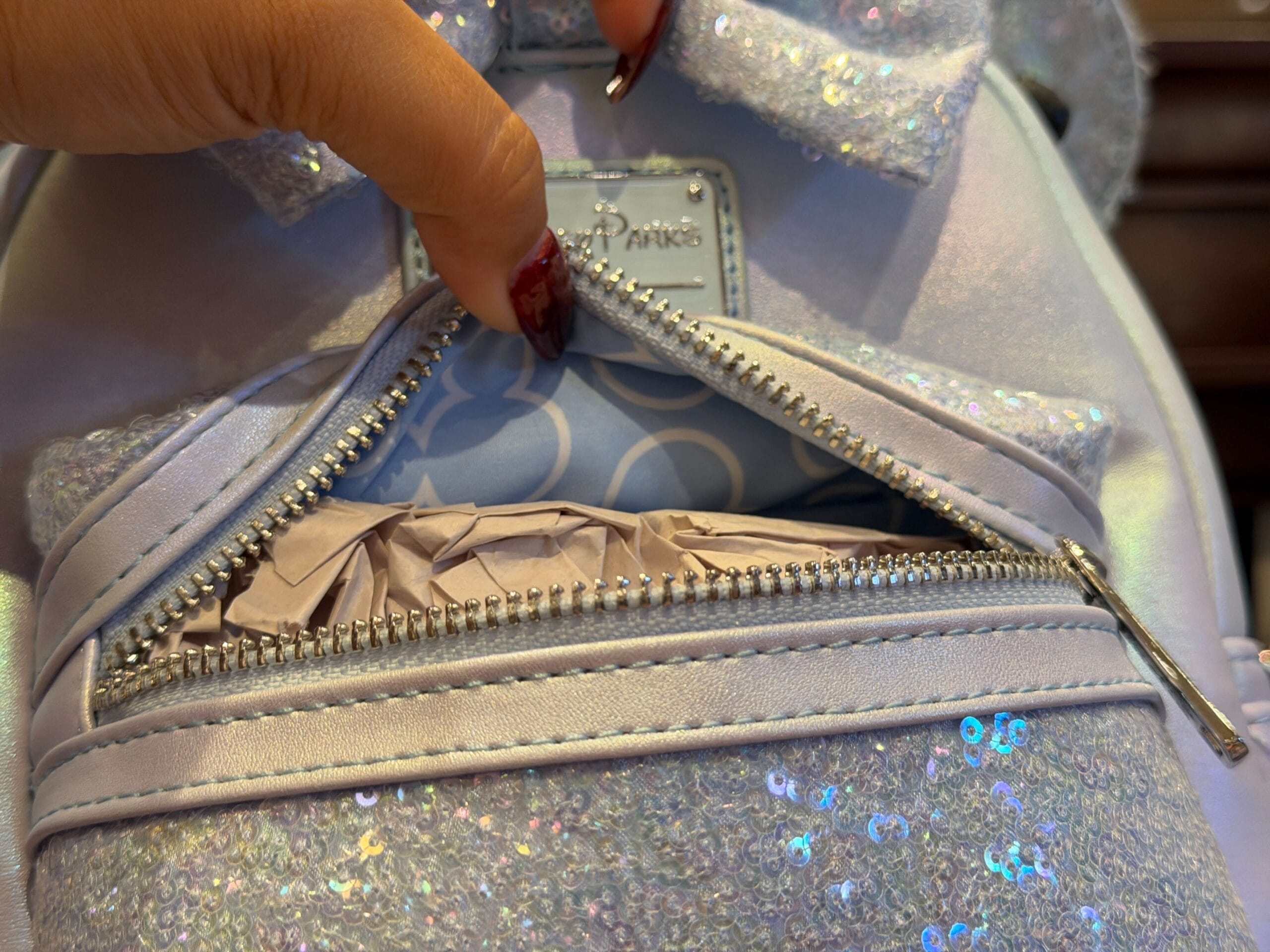 A hand opens a sparkly light blue bag, reminiscent of Cinderella Castle hues at Disney parks, revealing tissue paper inside the front pocket.
