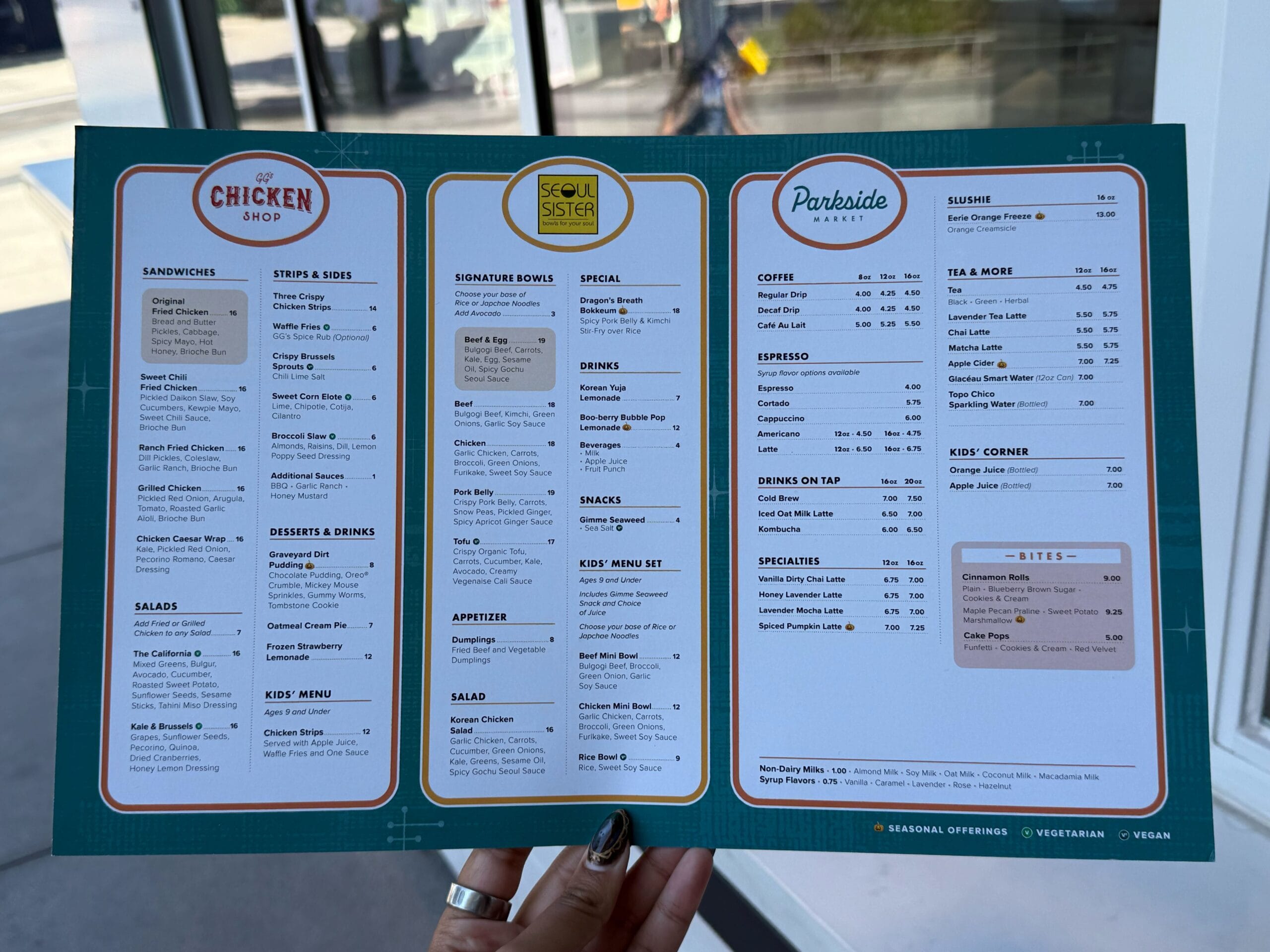 Blue menu board at a Disney park shows sections for sandwiches, sides, salads, bowls, coffee, tea, desserts, kids’ meals, and drinks.