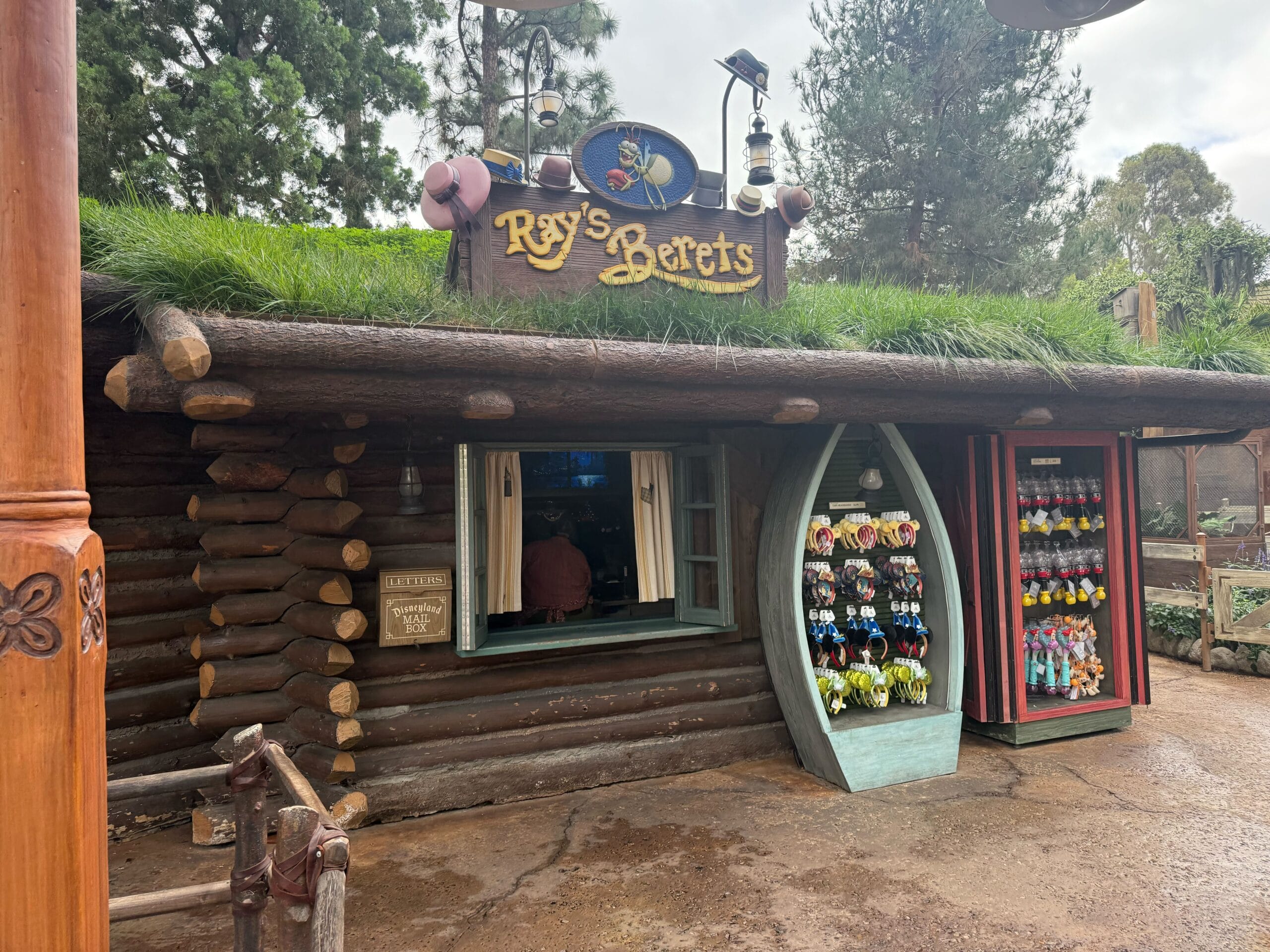 A log cabin shop named "Ray's Berets" sells hats and plush toys, its grassy roof giving a whimsical Disney village feel.