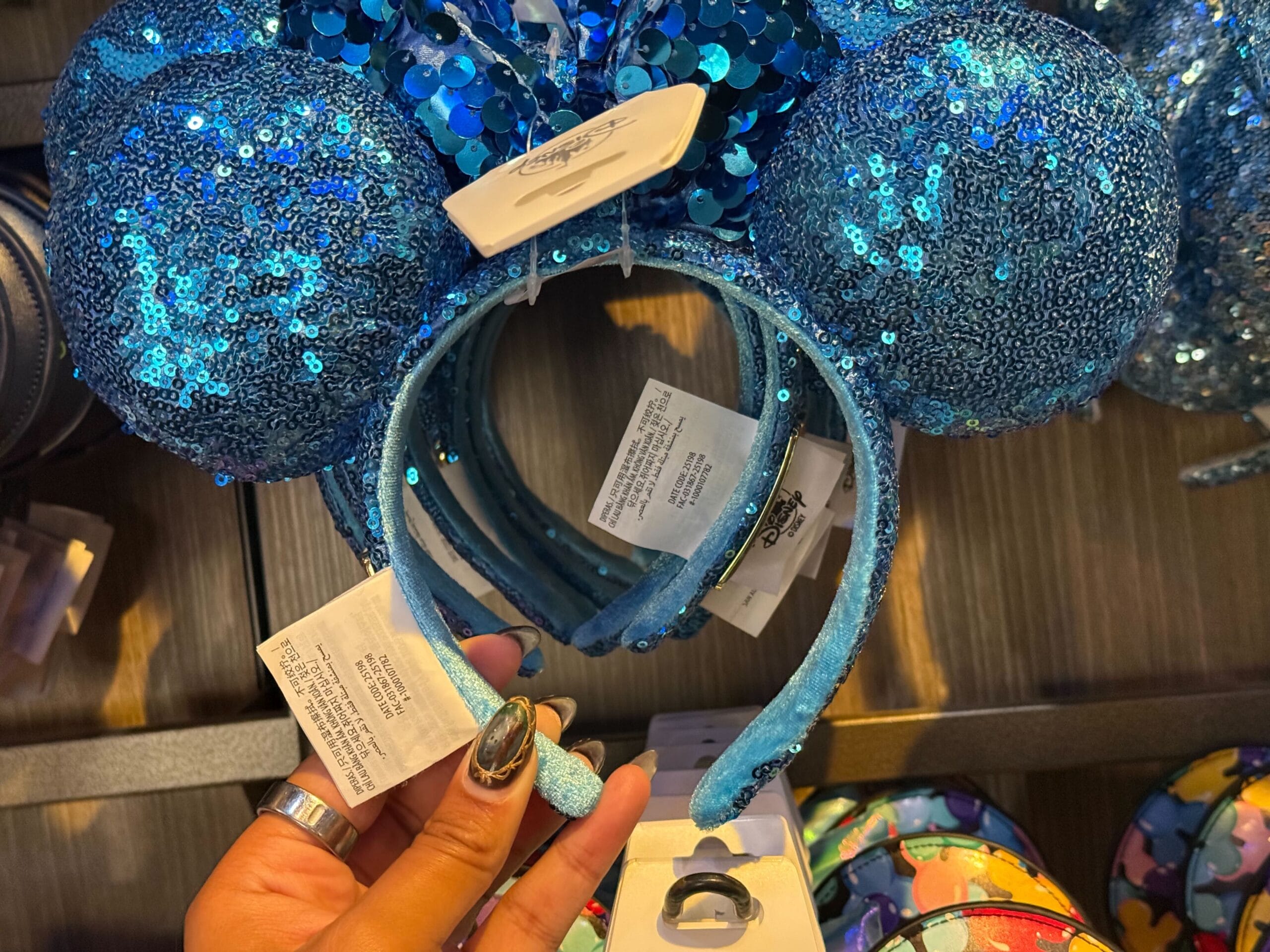A hand holds a blue sequined Mickey Mouse ear headband among similar styles in a Disney park store, shimmering with magic.