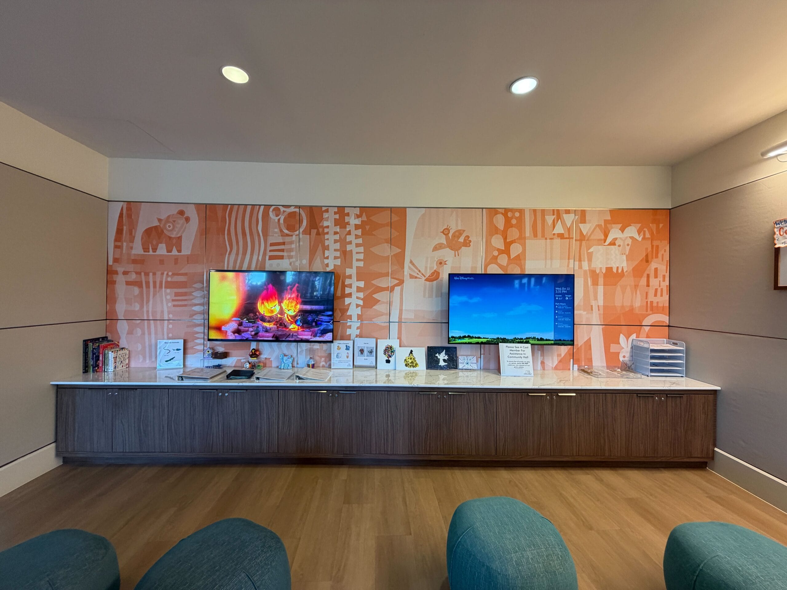 Two wall-mounted TVs above a wooden cabinet and decor on an orange mural wall at Bay Lake Tower’s modern Disney lounge.