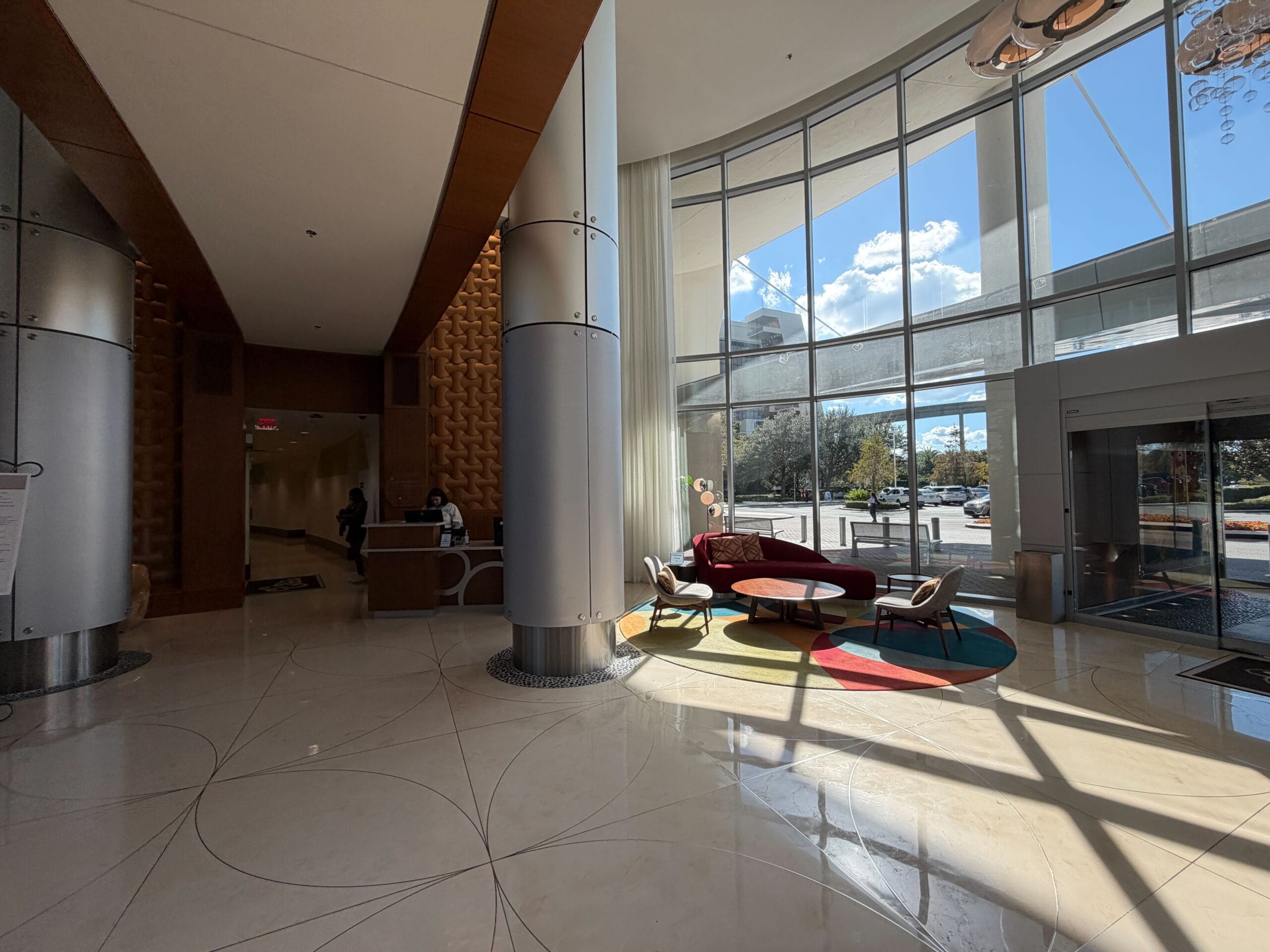 Modern Bay Lake Tower hotel lobby, Disney ambiance, sunlit windows, round rug, plush seating, and friendly Disney Cast Member.