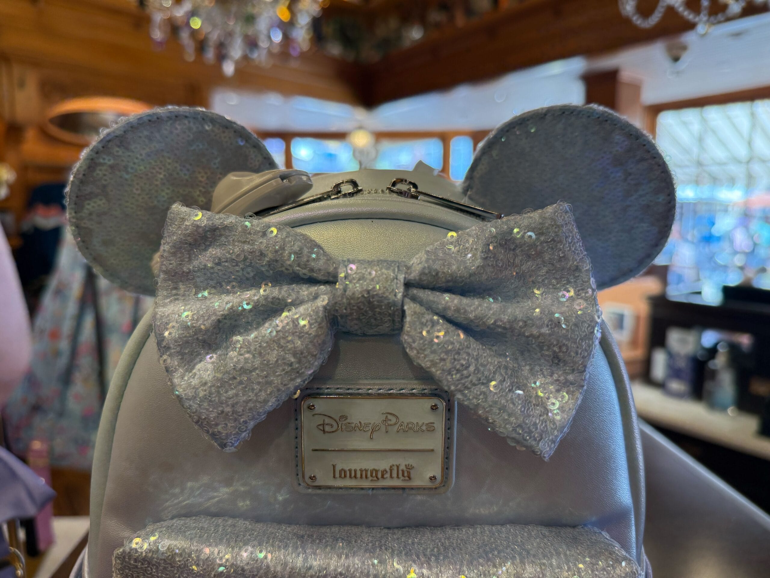 A Winter Blue Loungefly Backpack with silver sequins, mouse ears, and a bow displayed at Disneyland, near Sleeping Beauty Castle.