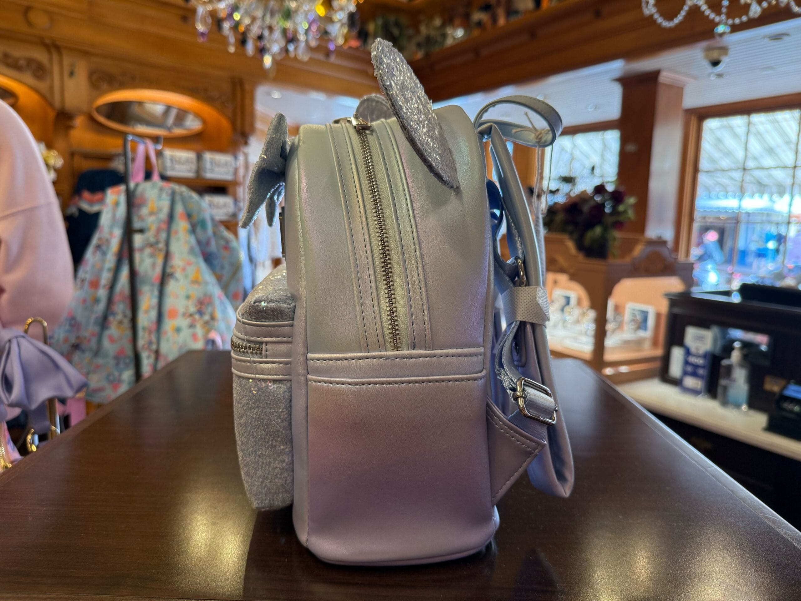 A pastel purple Disneyland backpack with sparkling mouse ears sits on a wooden store counter, evoking classic Disney park style.