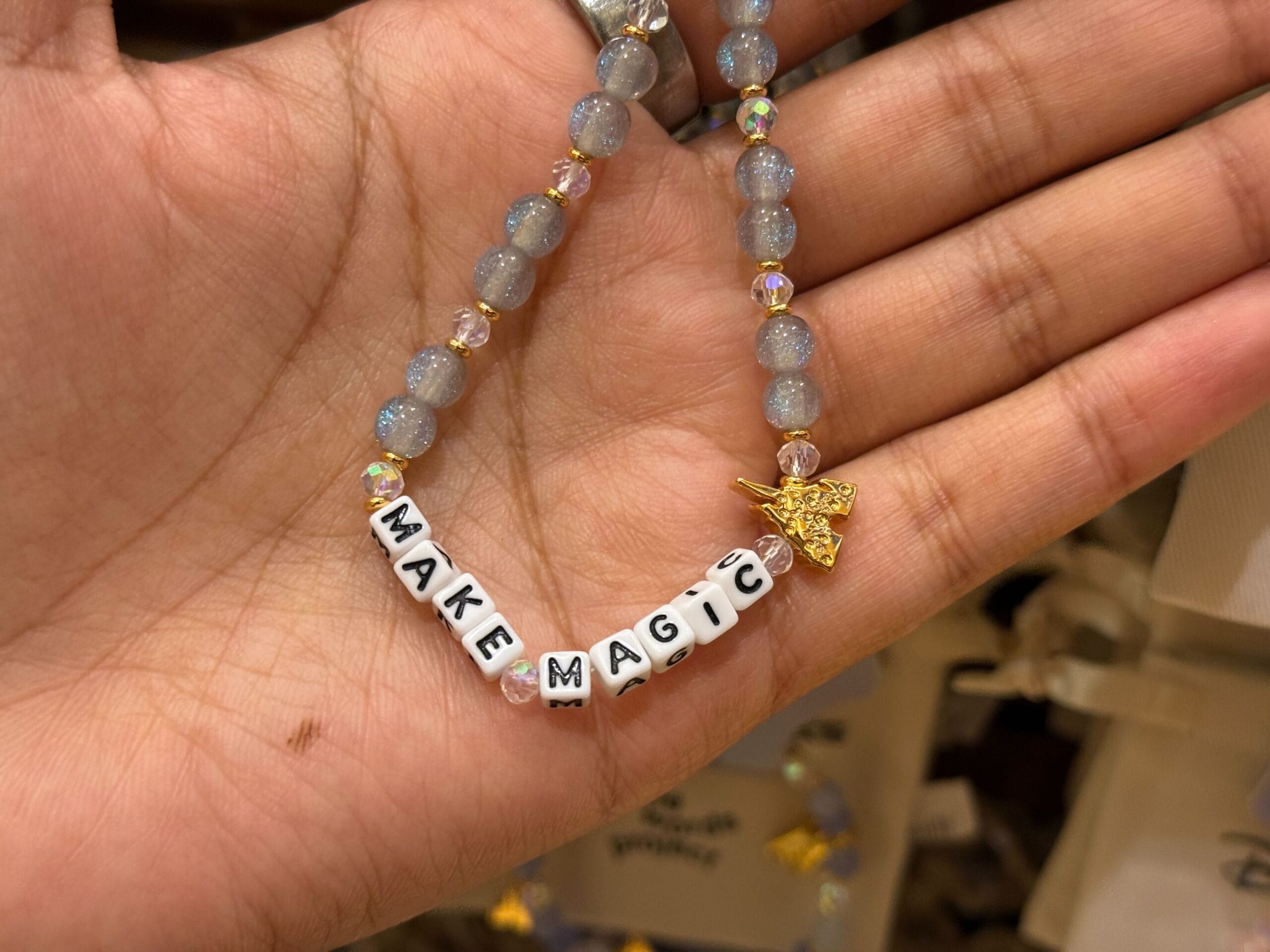 A hand at a Disney park holds a bracelet that spells "MAKE MAGIC" with a gold star charm, reminiscent of magical park moments.