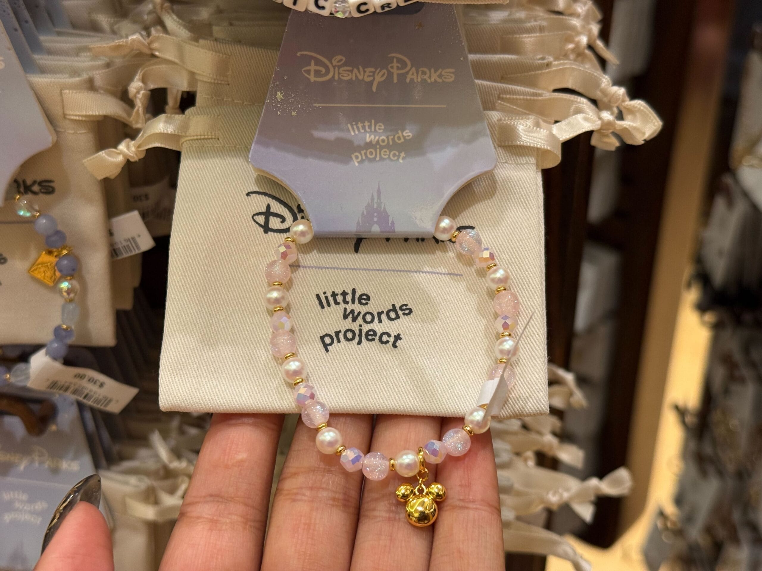 A hand displays a beaded bracelet with a gold Mickey charm and “little words project” tag at a Disney Parks shop display.
