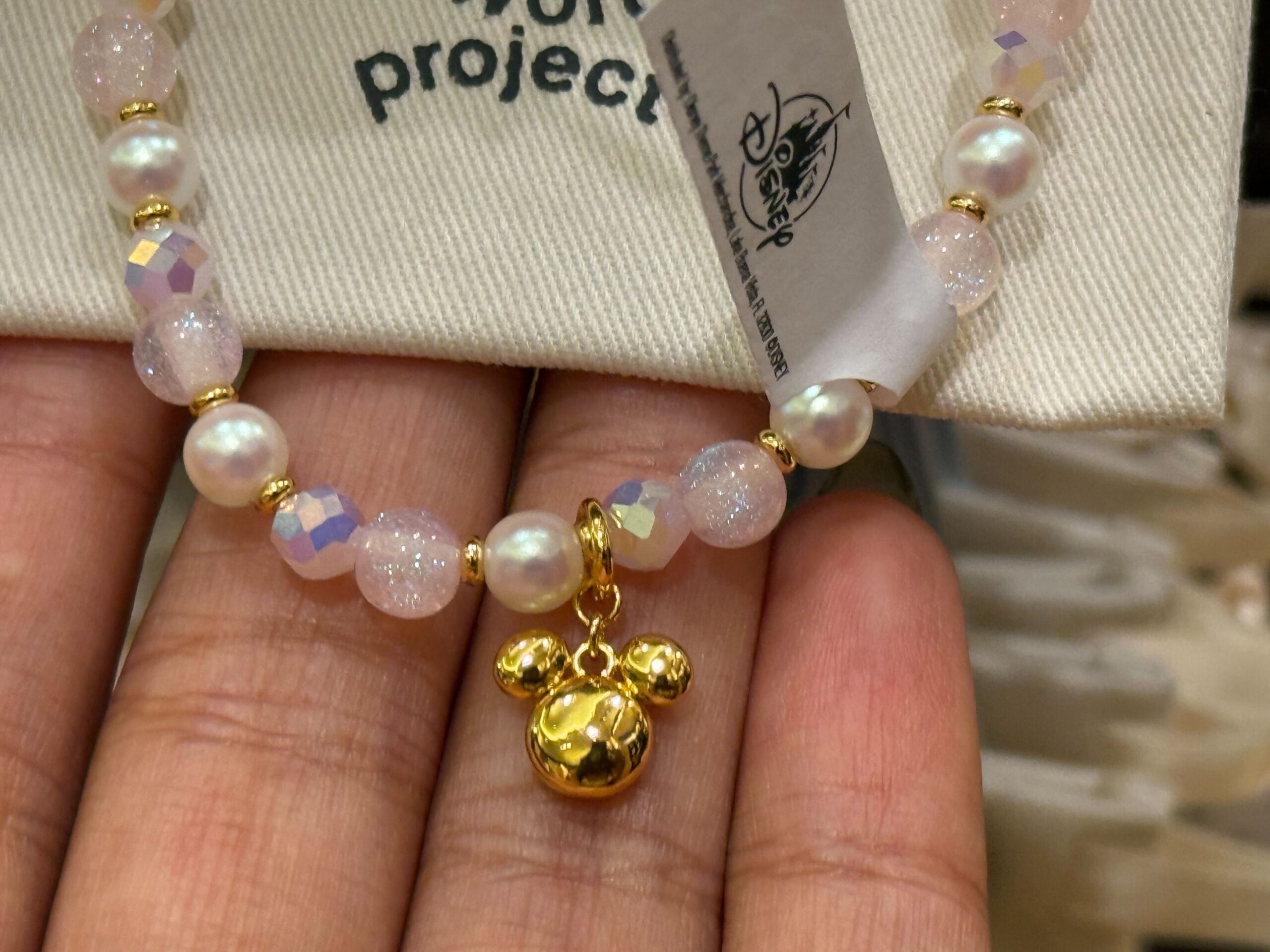 A beaded bracelet with a gold Mickey Mouse charm held between fingers, Disney tag visible—reminiscent of park souvenirs.