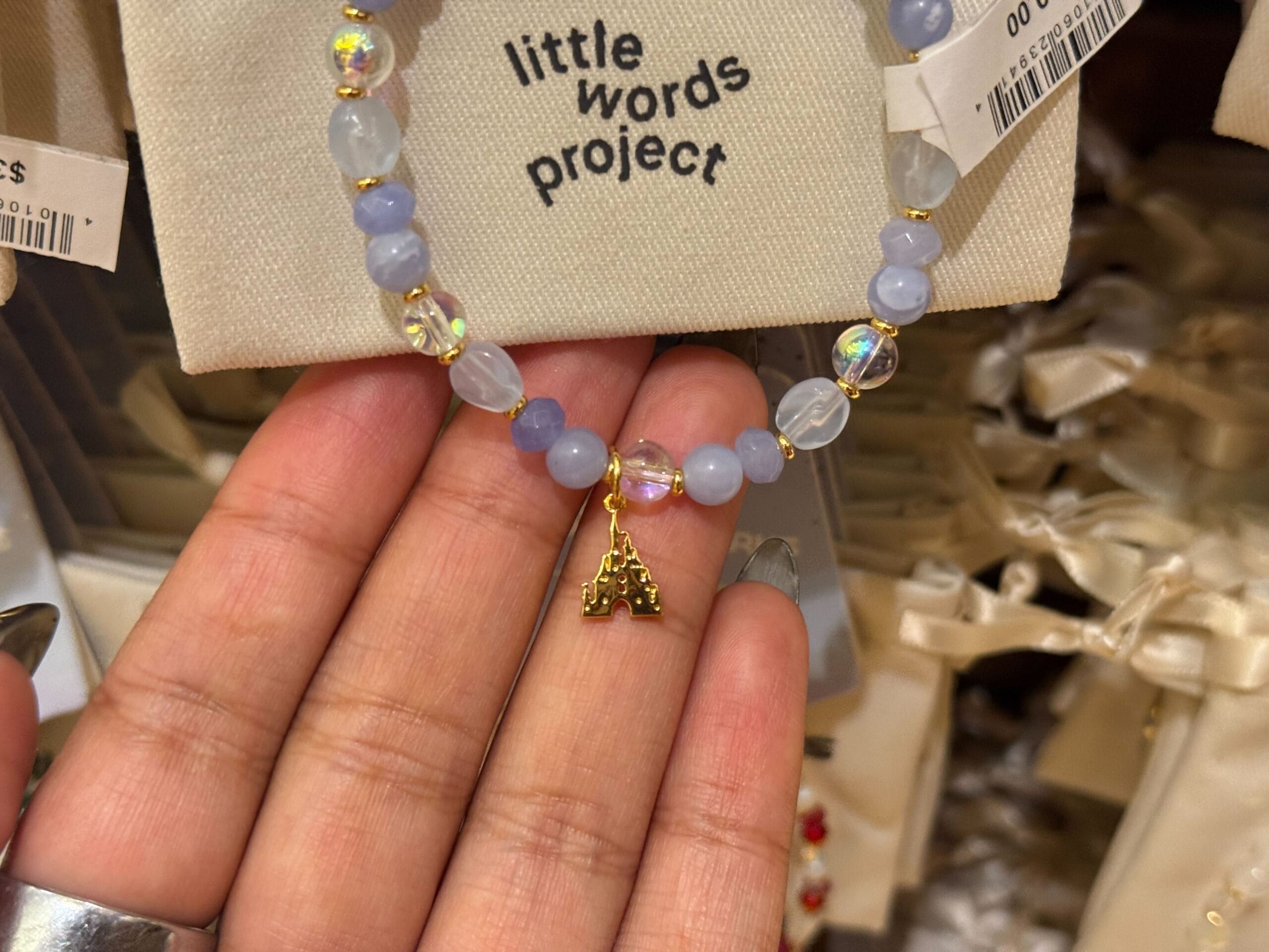 A hand holds a beaded bracelet with a gold "M" charm, "little words project" in the background—no Disney castle present.