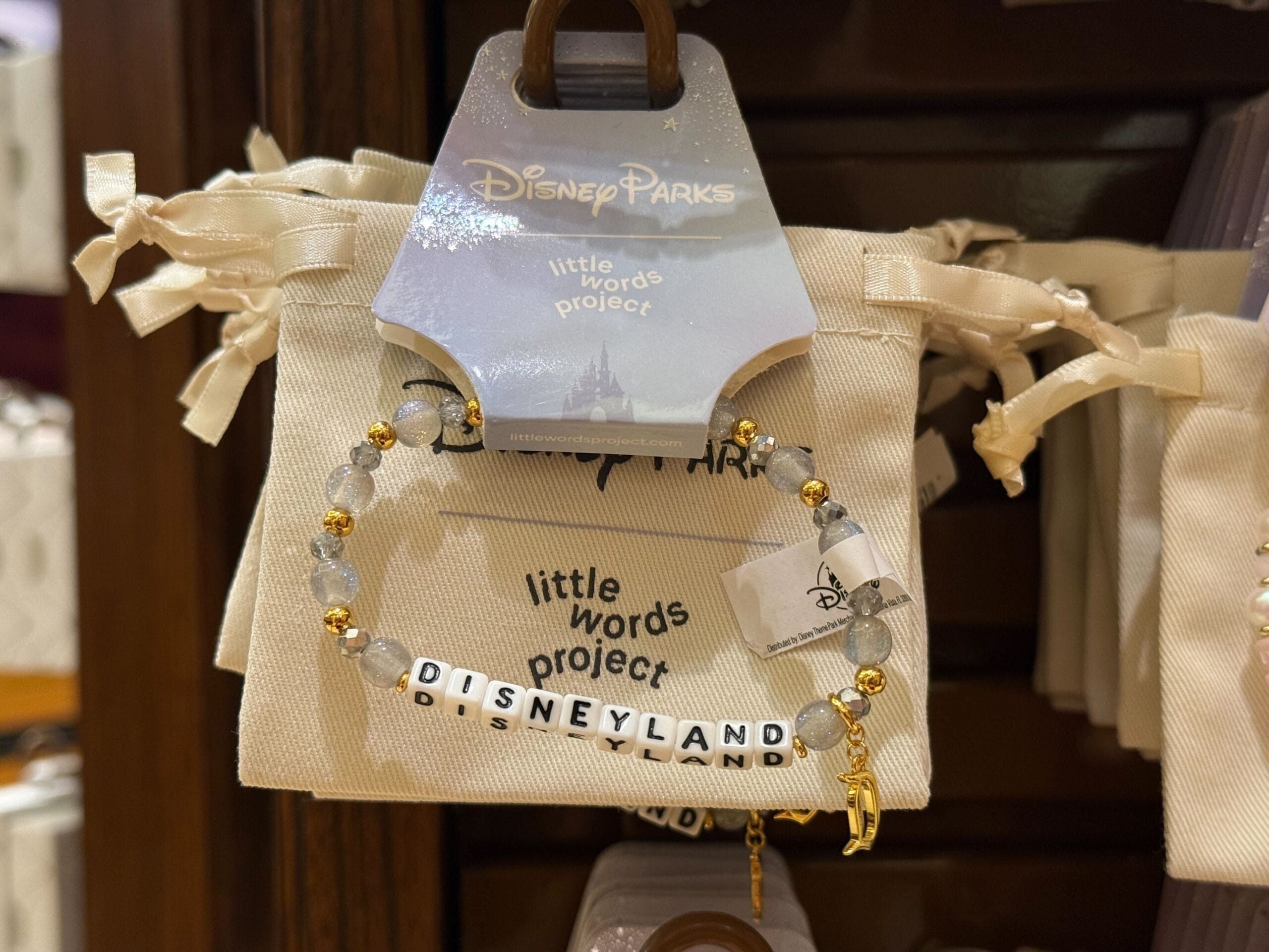 A beaded bracelet reading "DISNEYLAND" hangs on display, with Disney Parks packaging reminiscent of worldwide park souvenirs behind it.