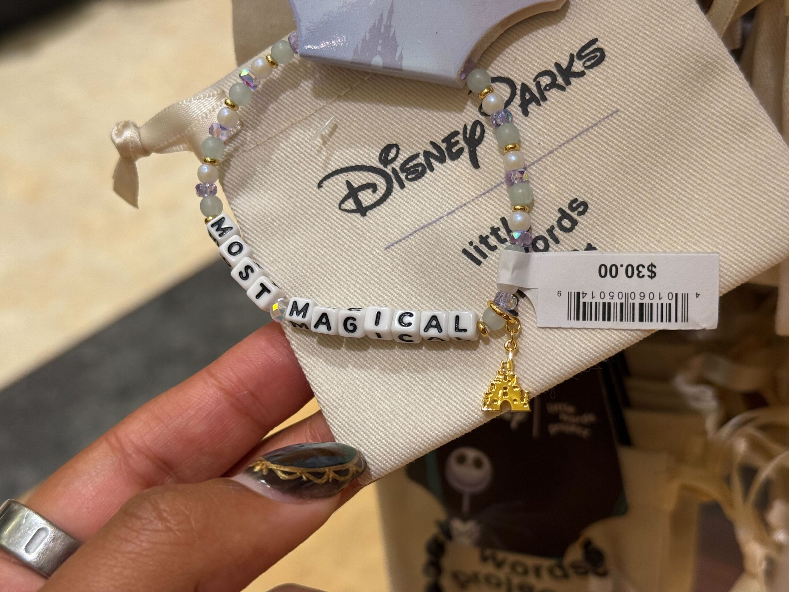 A hand displays a "MOST MAGICAL" beaded bracelet with a gold castle charm, similar to iconic Disney Park castles, $30 at Disney Parks.