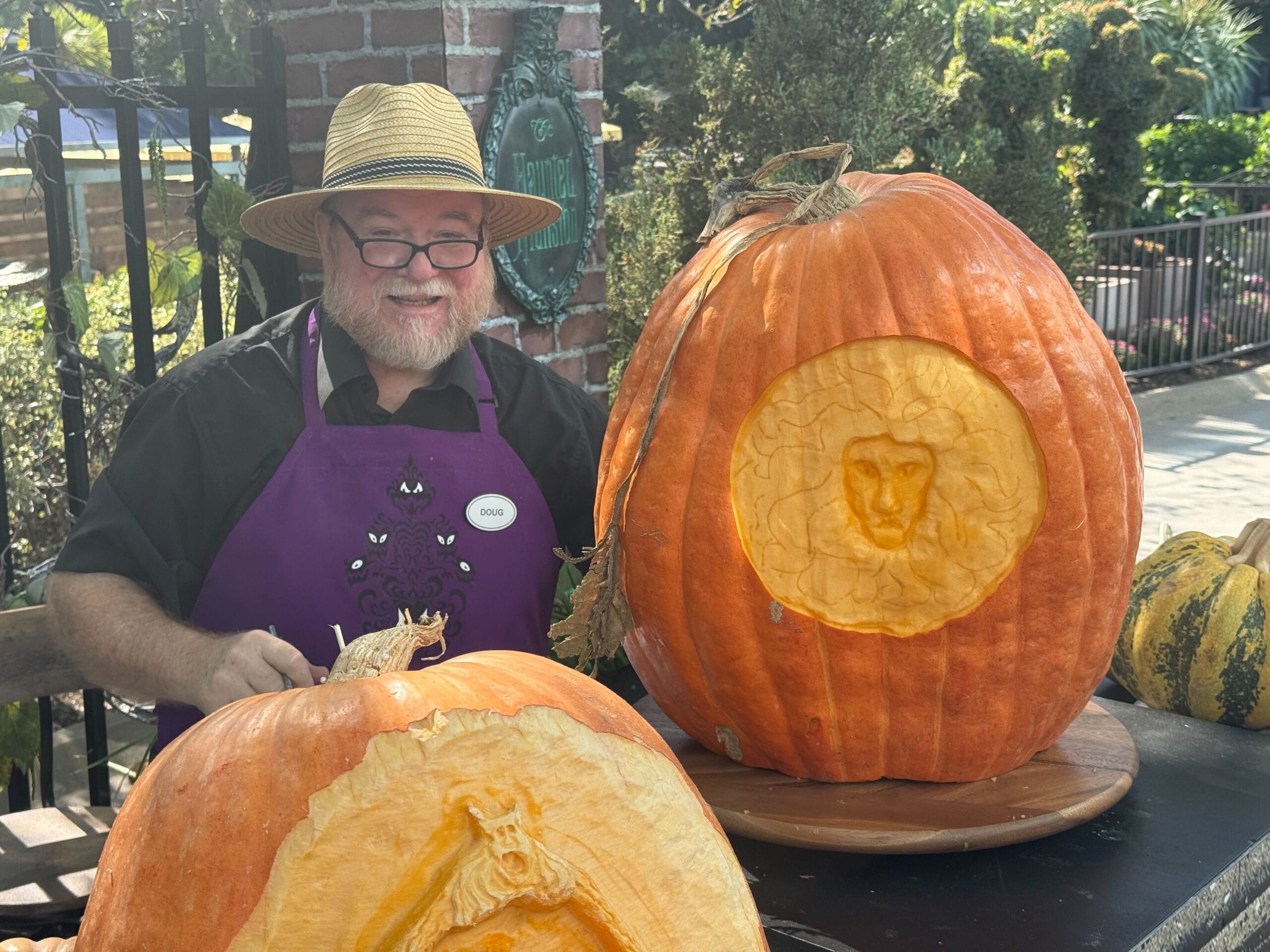 A man in a straw hat and purple apron sits beside two large pumpkins carved with detailed lion faces, evoking Disney’s artistry.