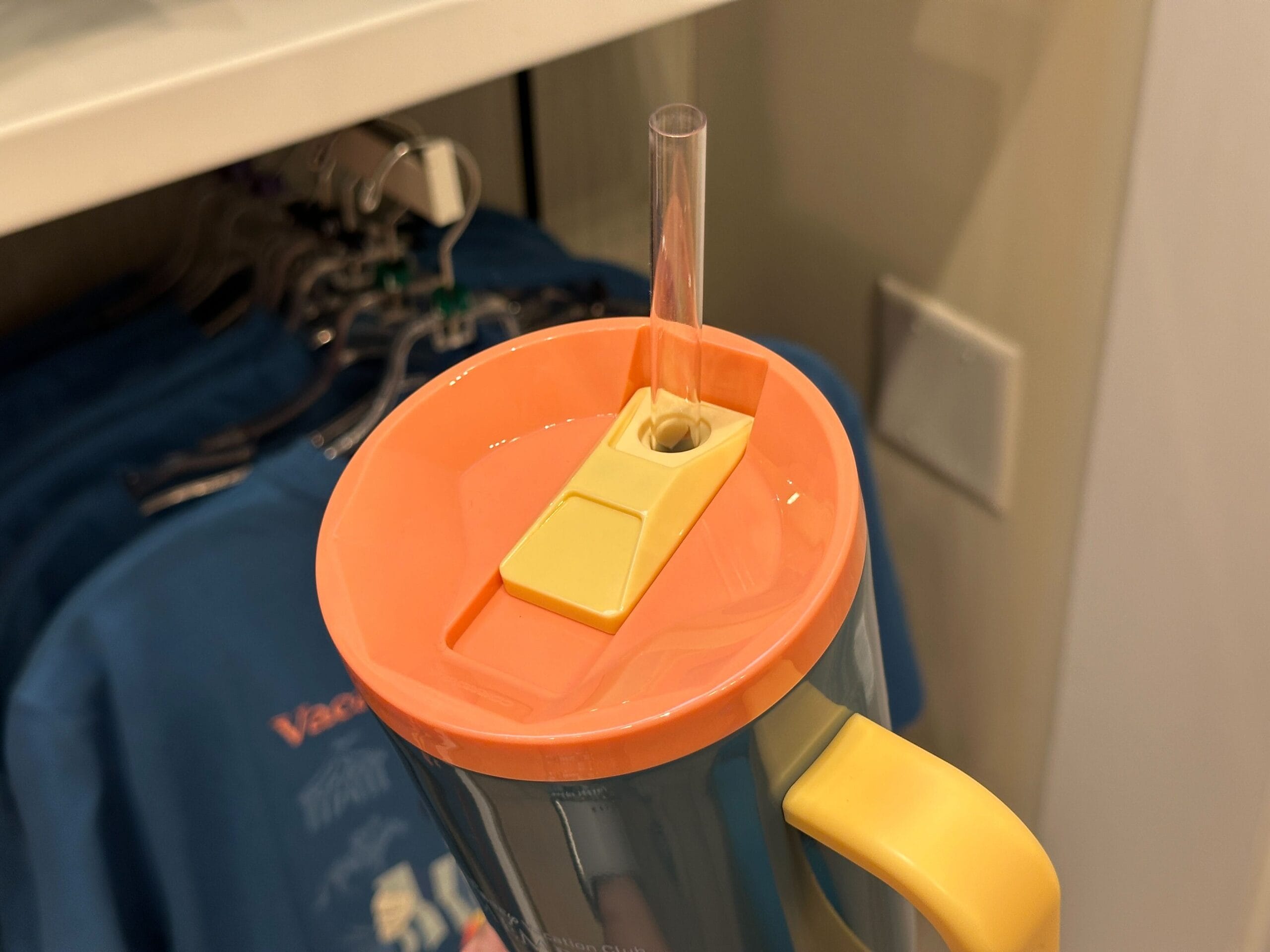 A close-up of a cup with an orange lid and clear straw. Grand Floridian blue shirts from the new collection hang in the background.