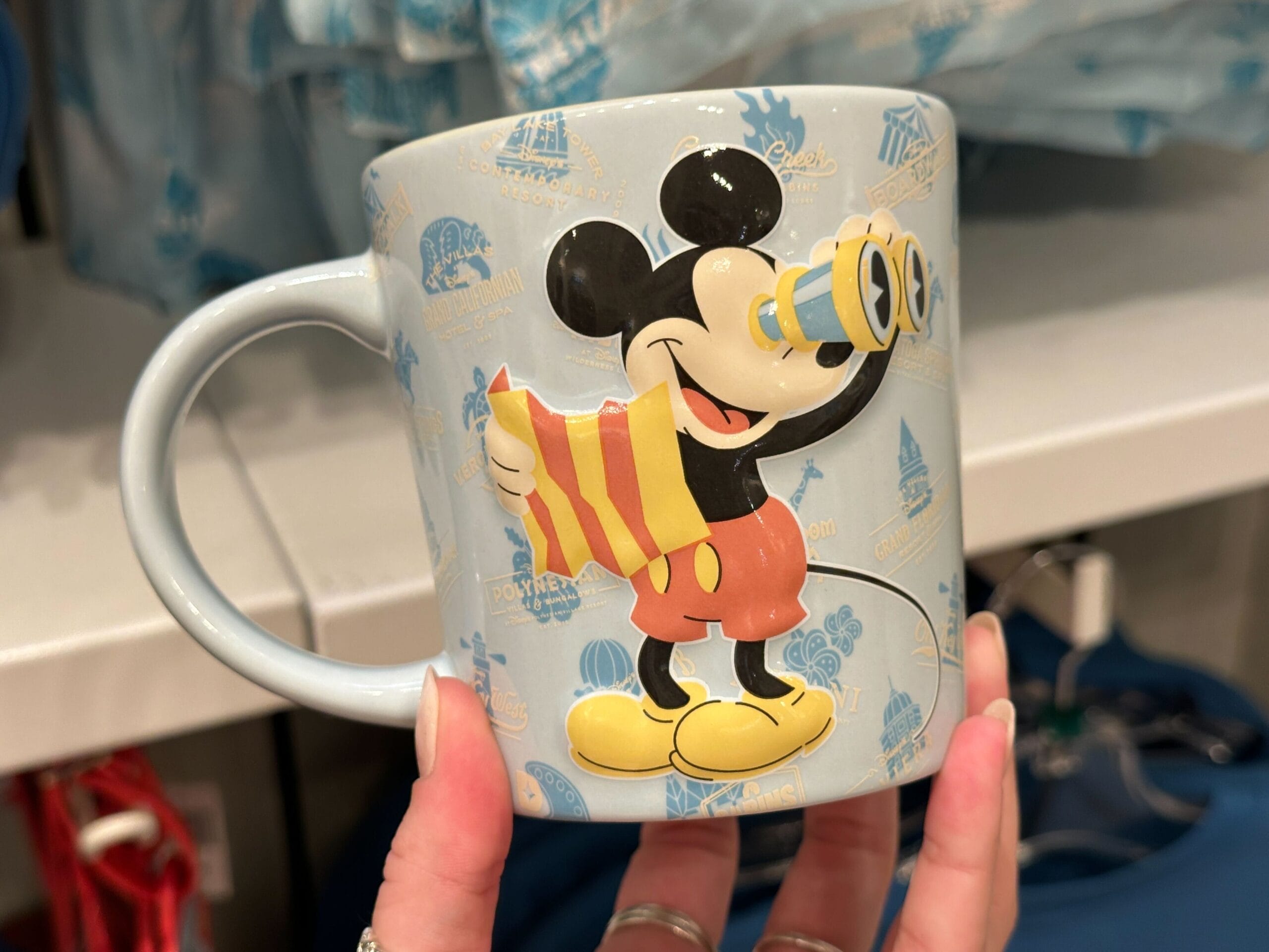A hand holds a mug from the new collection, featuring Mickey Mouse with a map and binoculars.