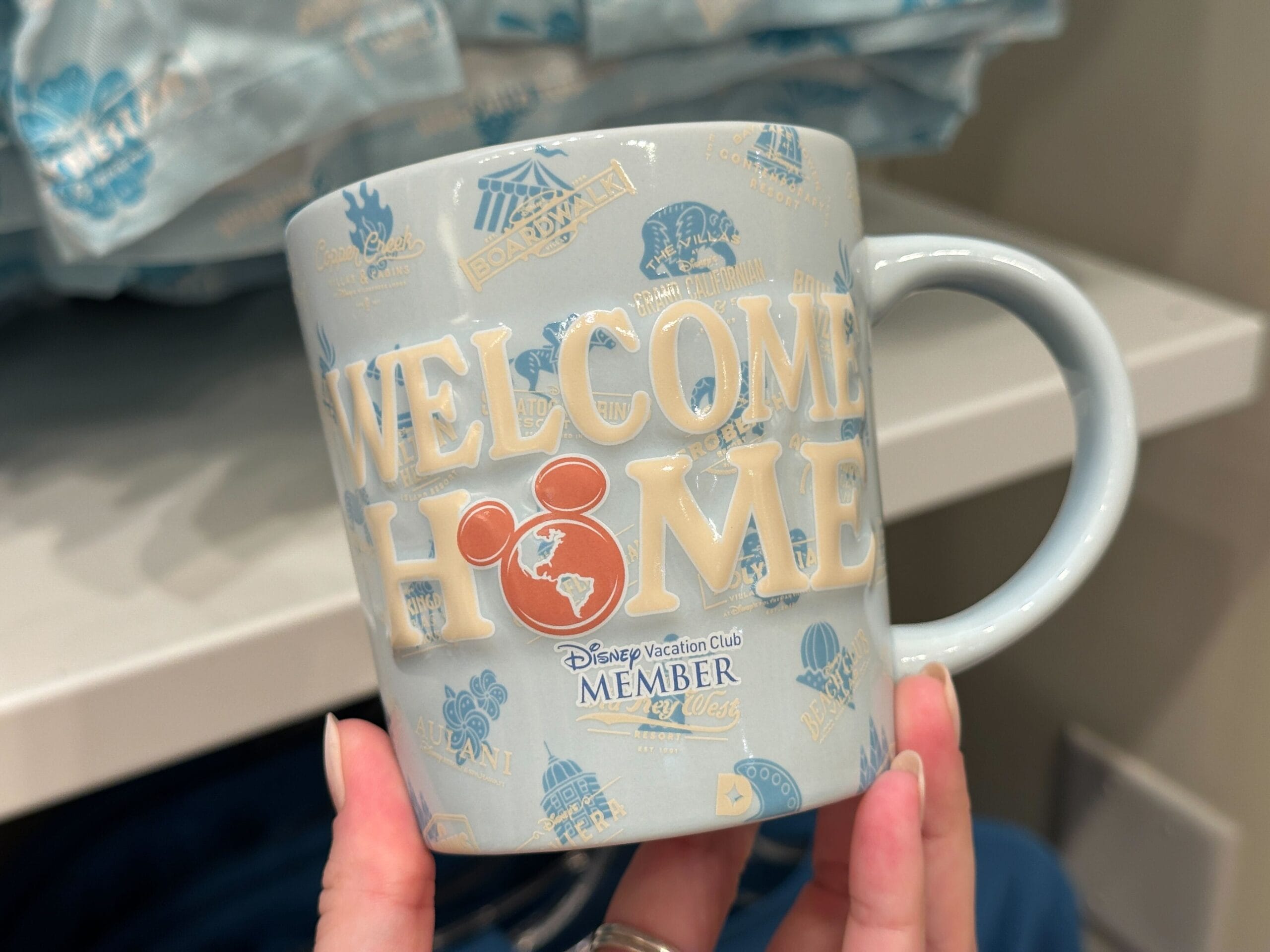 A hand holds Grand Floridian DVC Member Merch: a mug with Disney icons and "Welcome Home" message.
