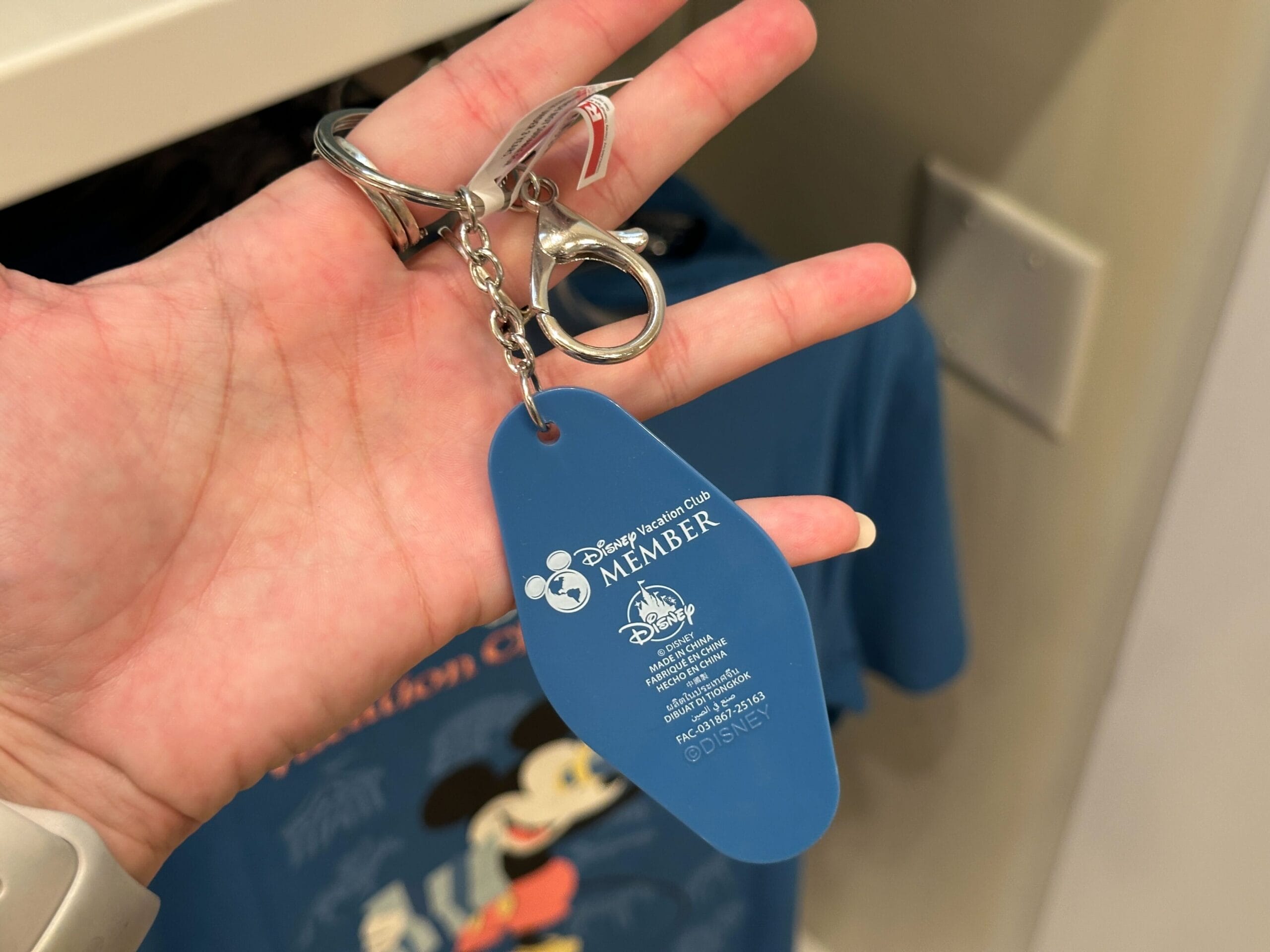 A hand holds a blue DVC Member Merch keychain in front of a blue Mickey Mouse shirt.