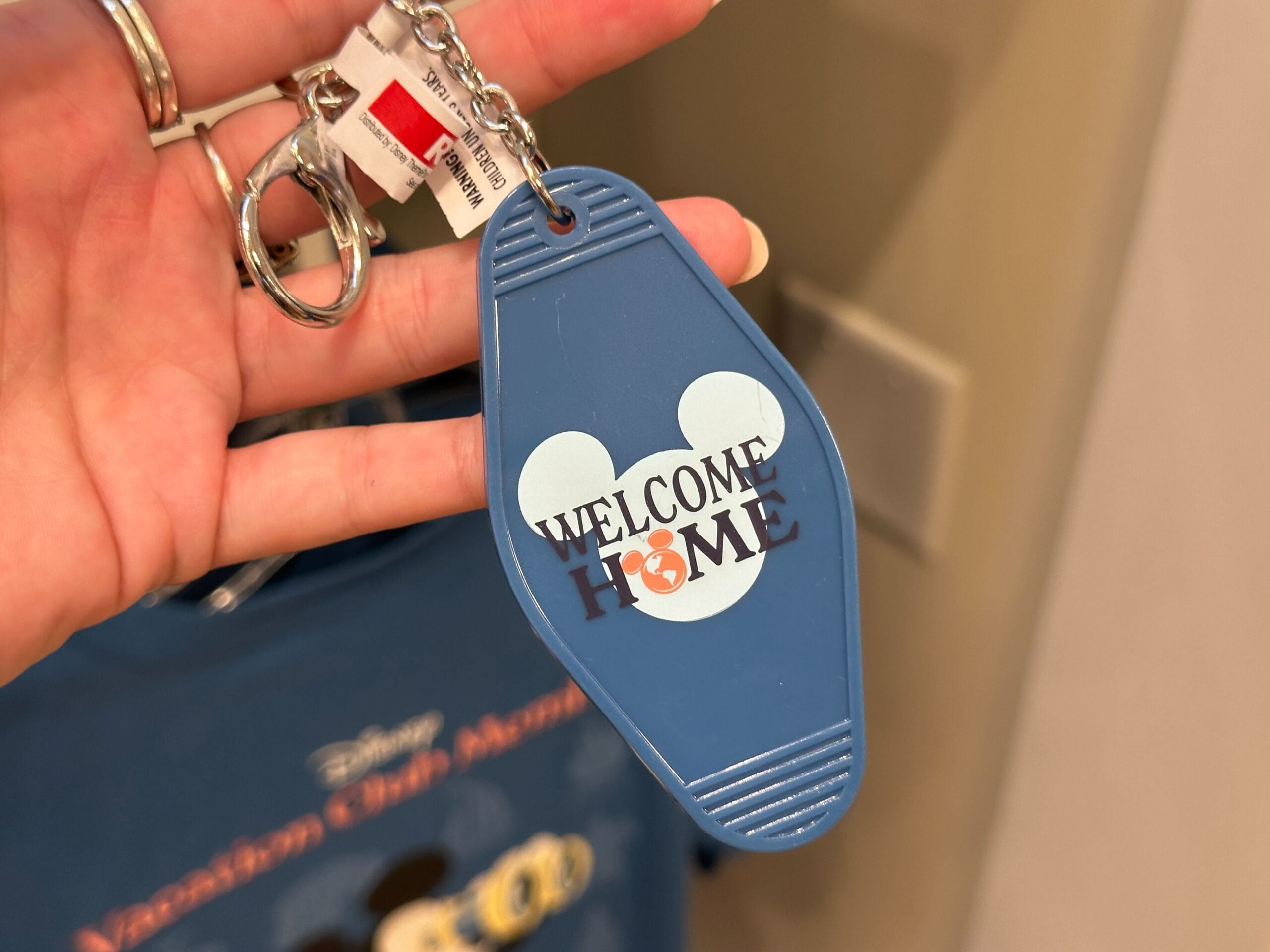 A hand holds a blue DVC Member keychain with a Mickey Mouse head and "WELCOME HOME" printed on it.
