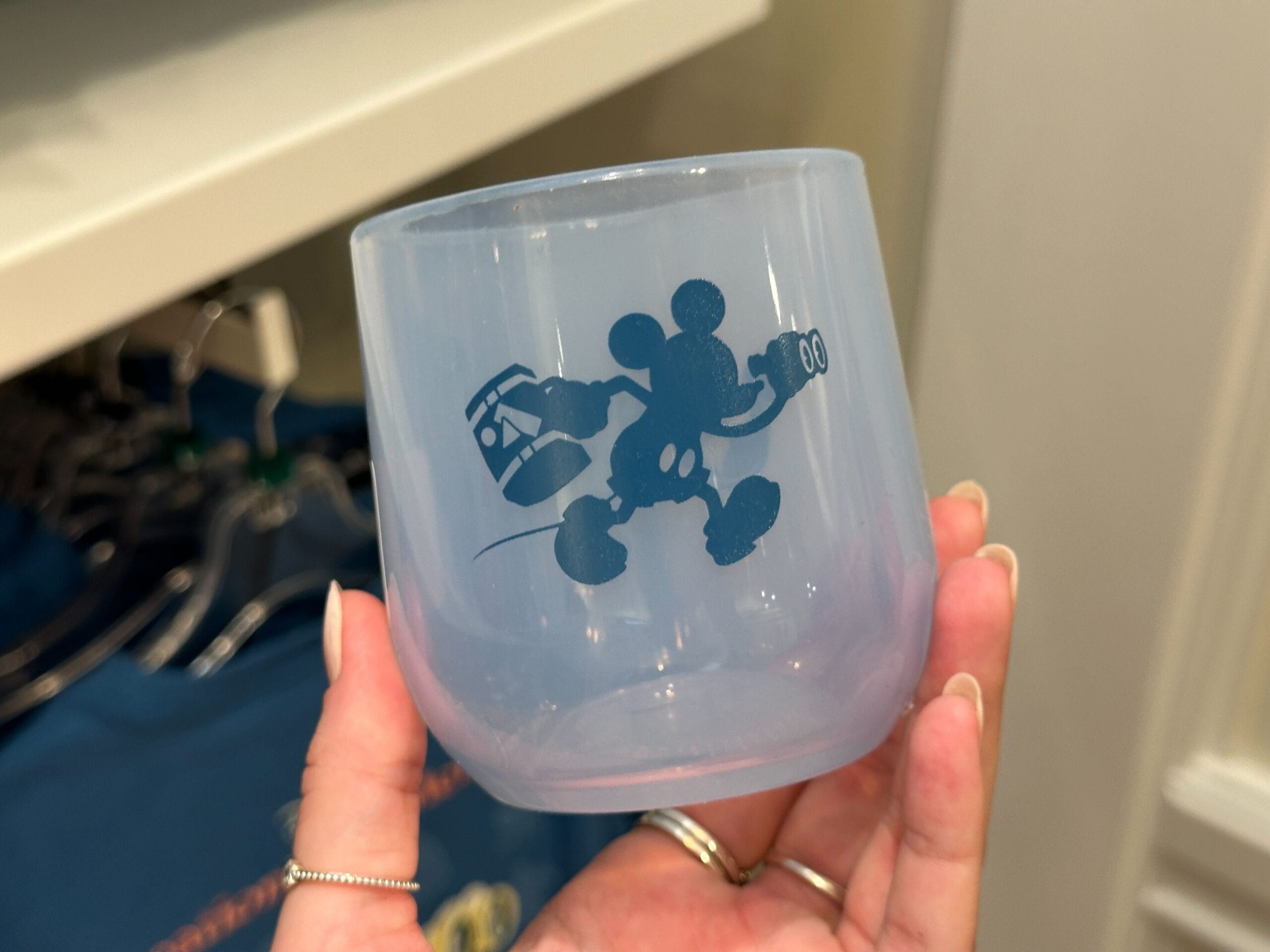 A hand holds a translucent cup from the Grand Floridian Merch Collection, featuring Mickey Mouse with a lantern and backpack.