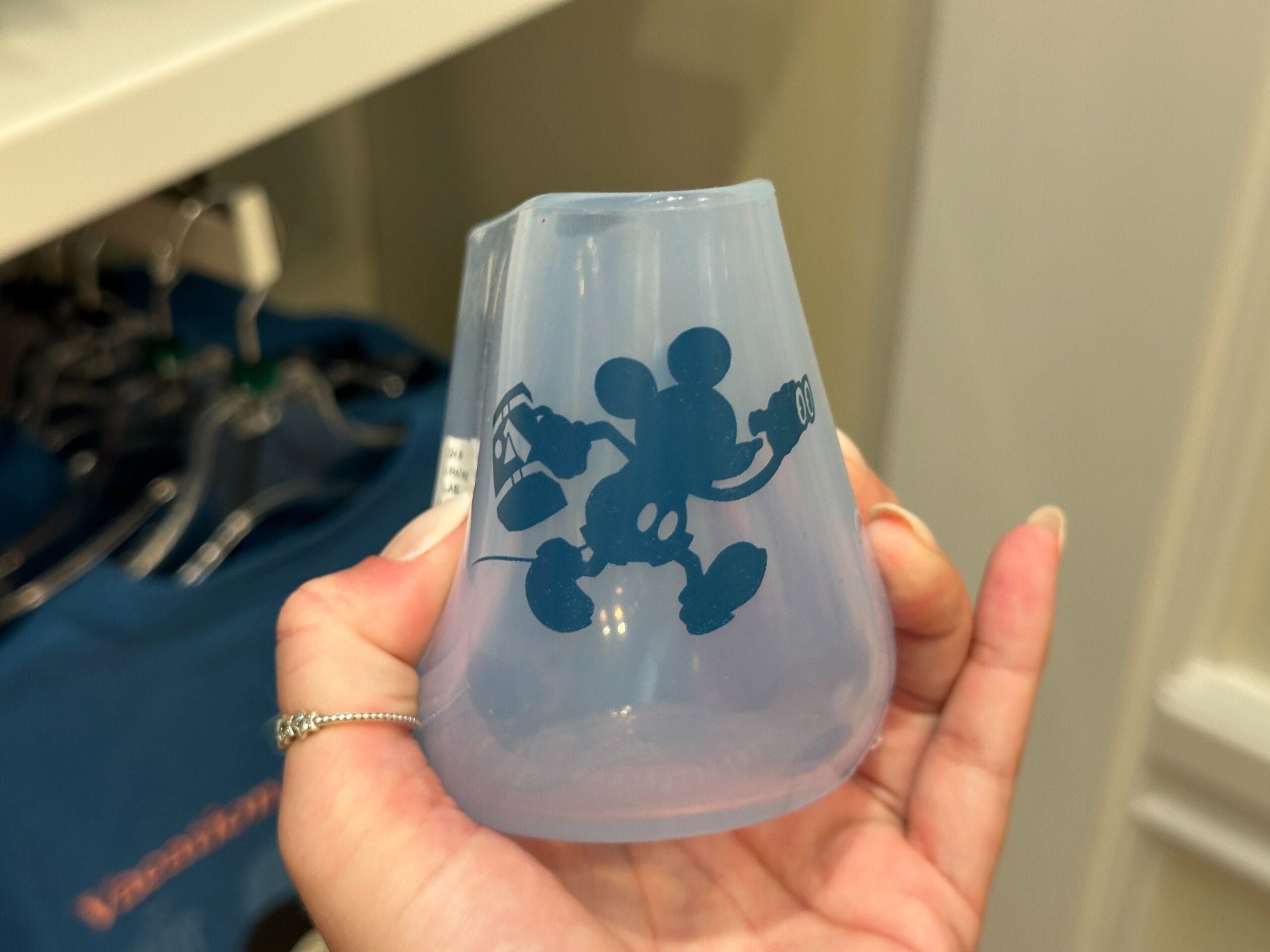 A hand holds a small translucent cup from the DVC Member Merch New Collection, featuring Mickey Mouse with a camera.