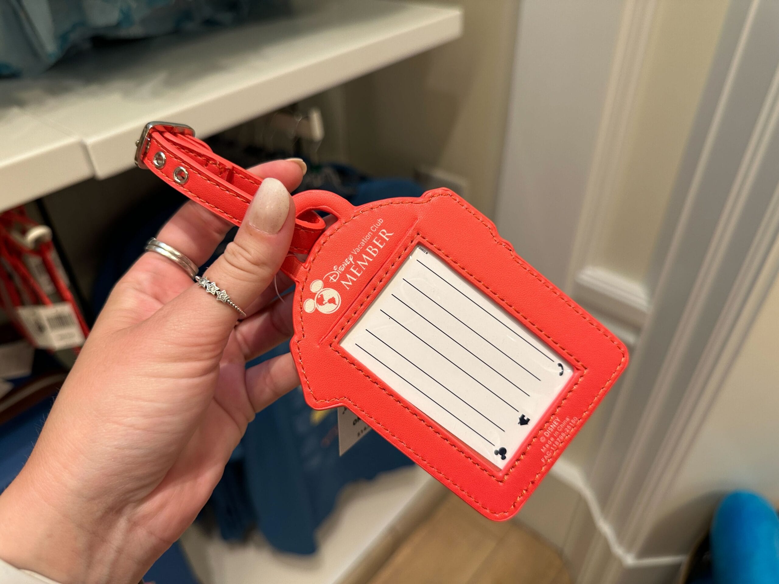 A hand holds a red Disney-themed luggage tag from the DVC Member Merch New Collection with blank lines for info.