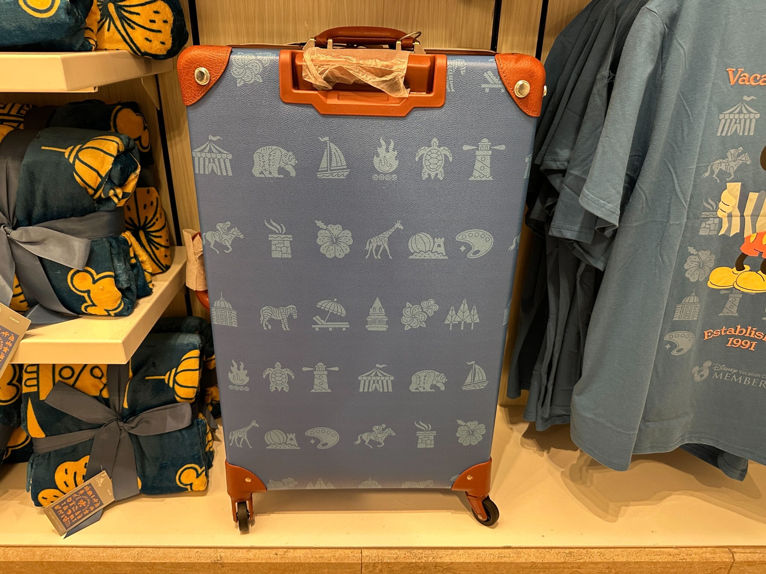Blue suitcase with tan accents from the Grand Floridian merch collection, featuring white icons, displayed by blue shirts.
