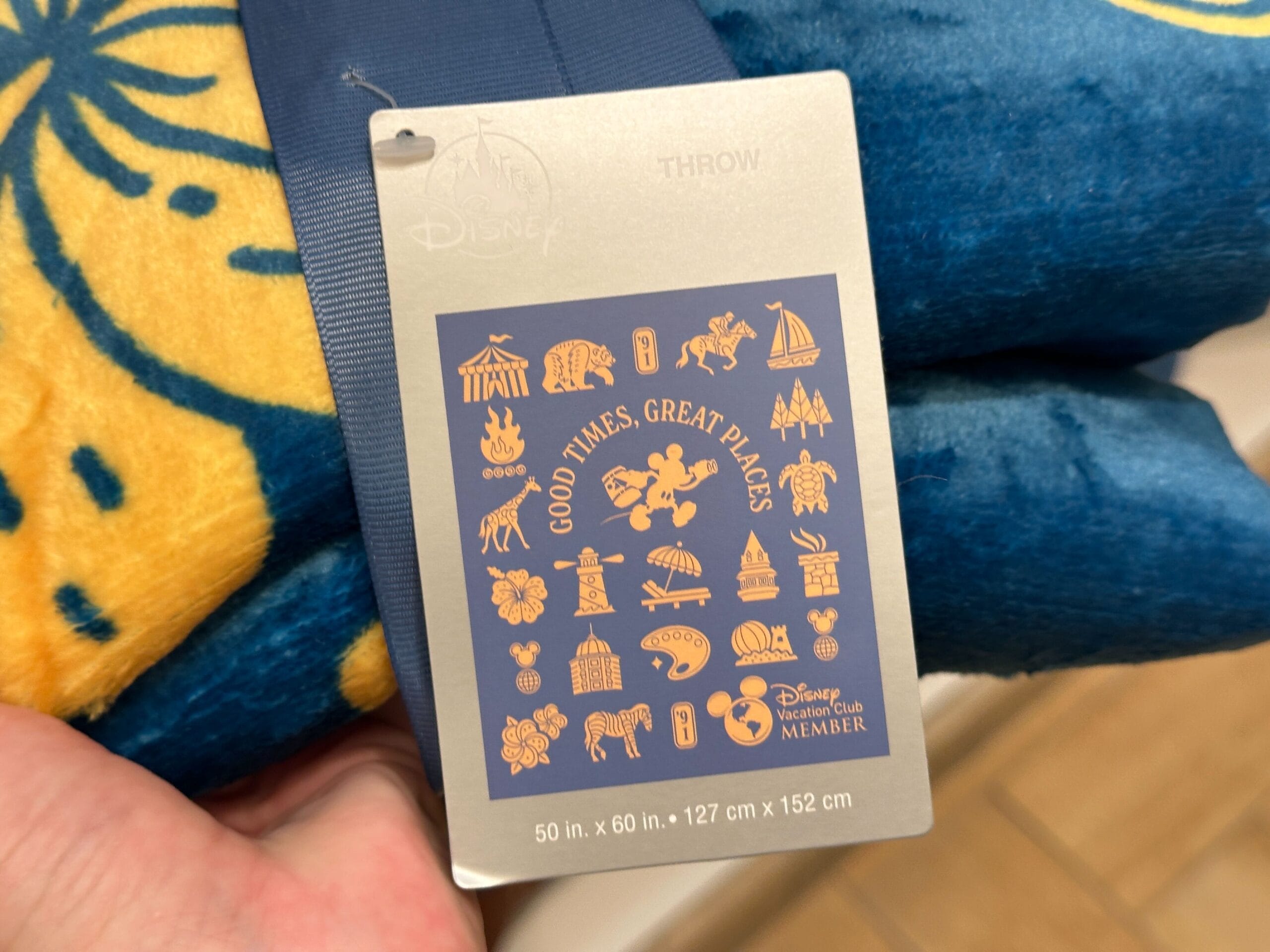 A Disney throw blanket tag from the New Collection with icons and “Good Times, Great Places” in gold on blue.